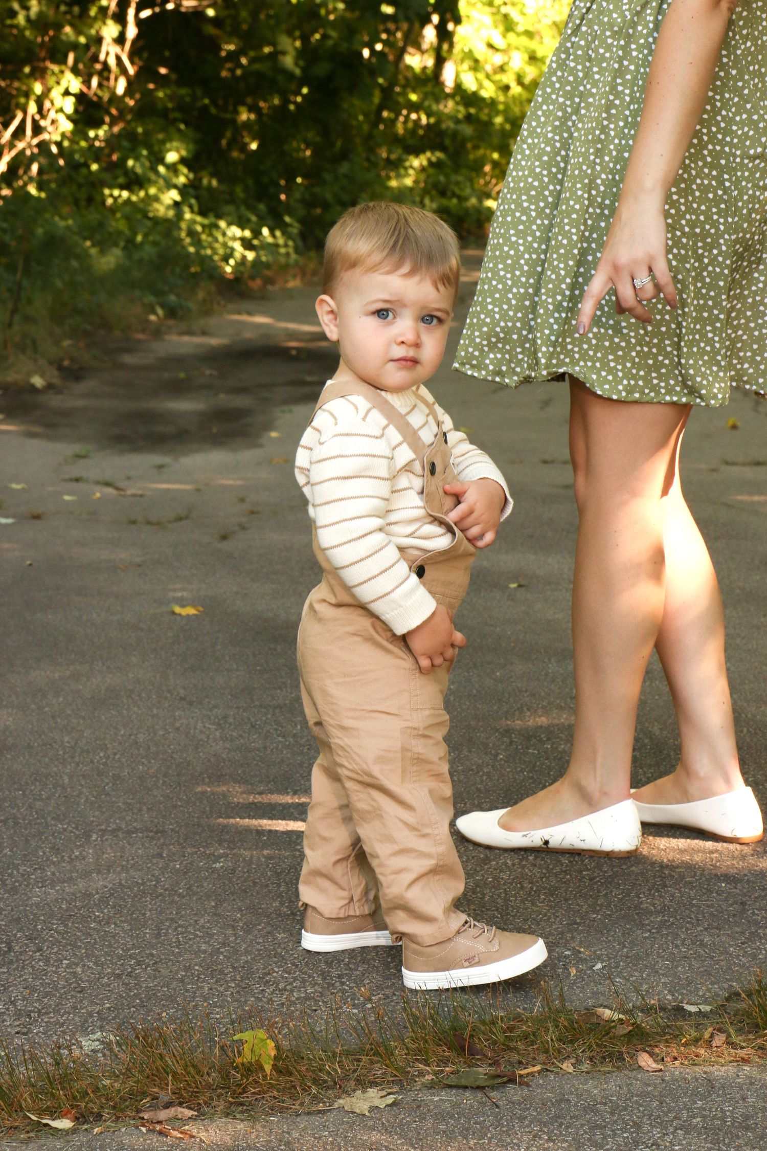 Adorable toddler boy in tan overalls and striped shirt standing on path beside mother outdoors.