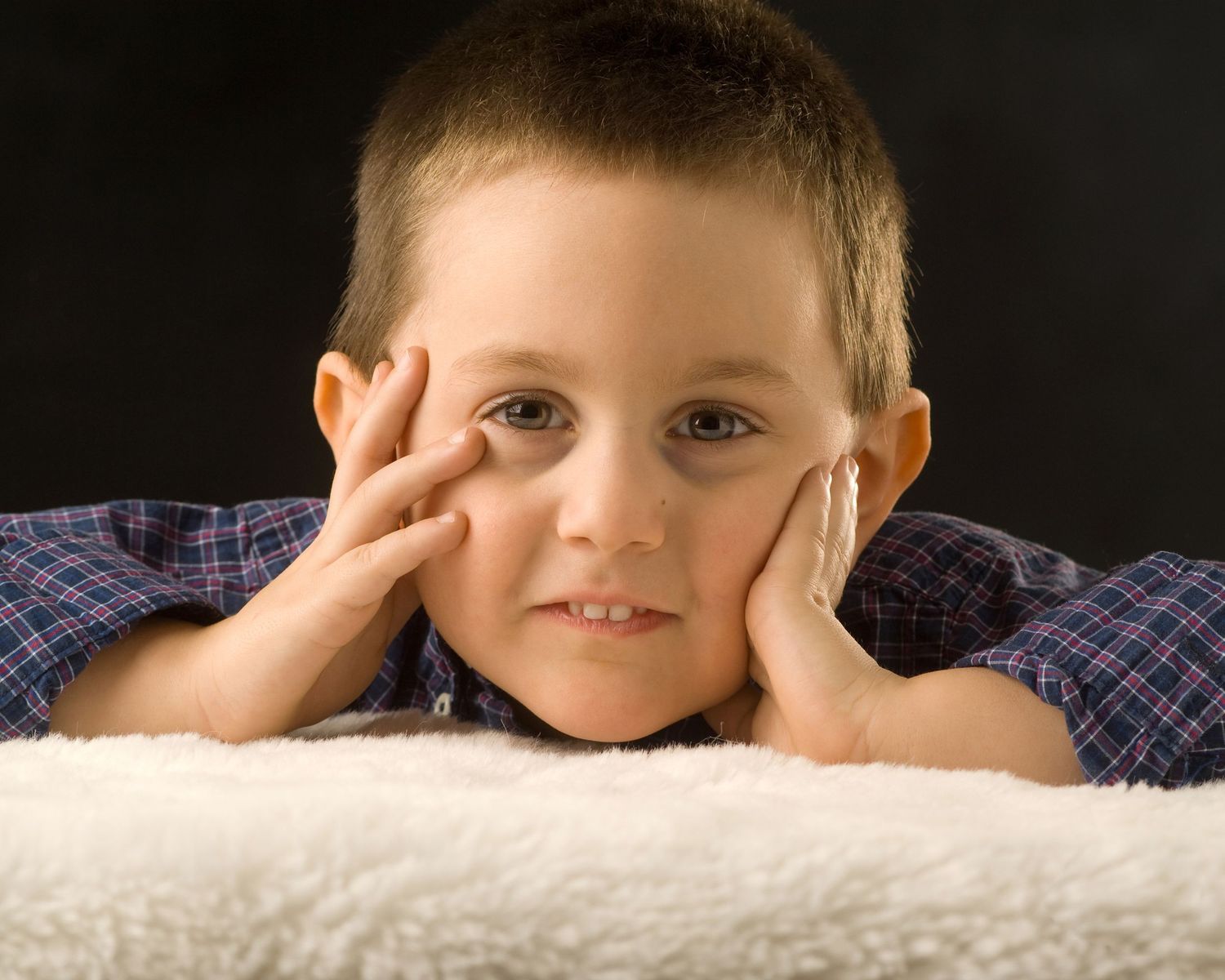 Cute young boy resting chin on hands with thoughtful expression against dark background.