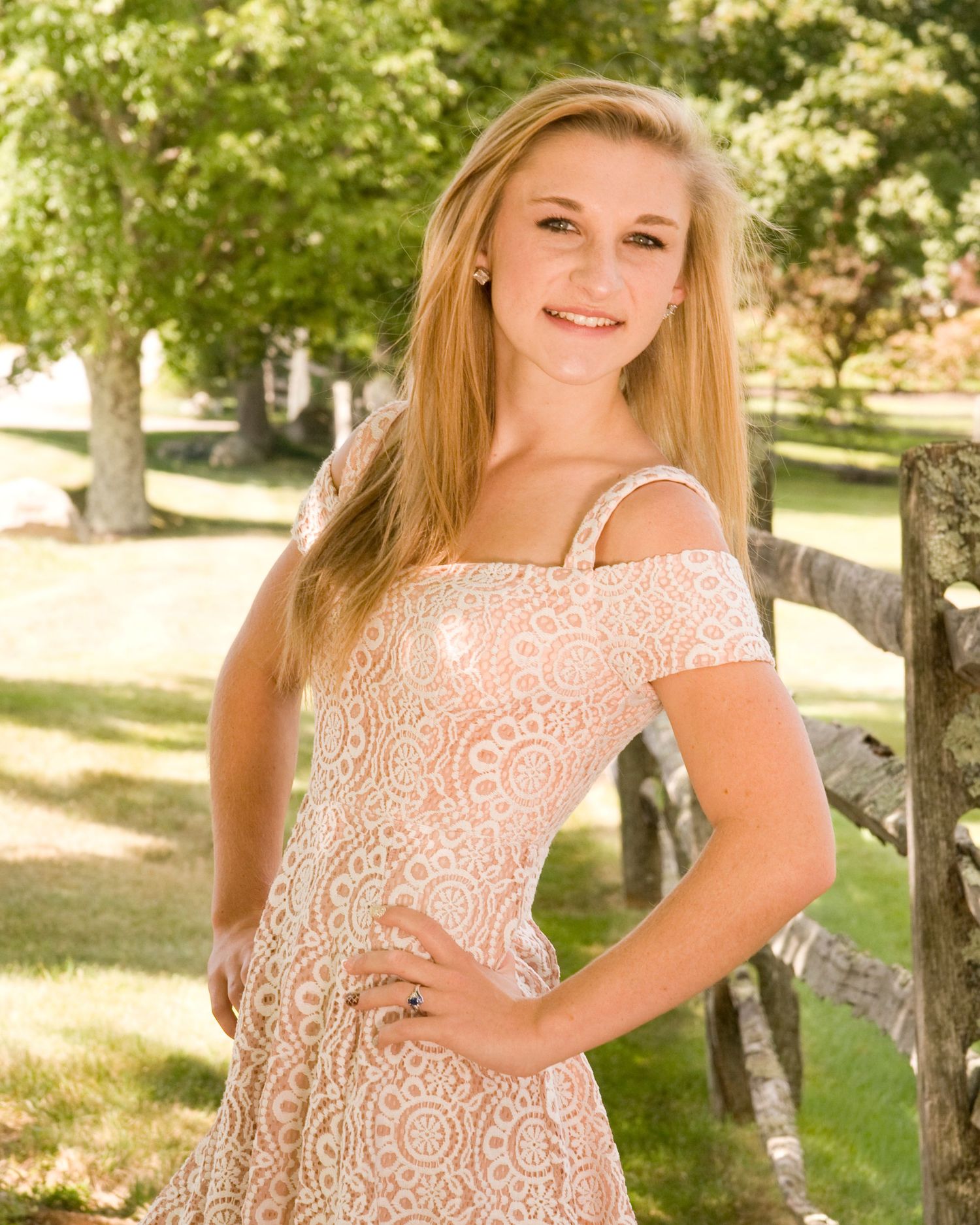 Young blonde woman in lace off-shoulder dress poses by rustic fence in sunny outdoor setting.