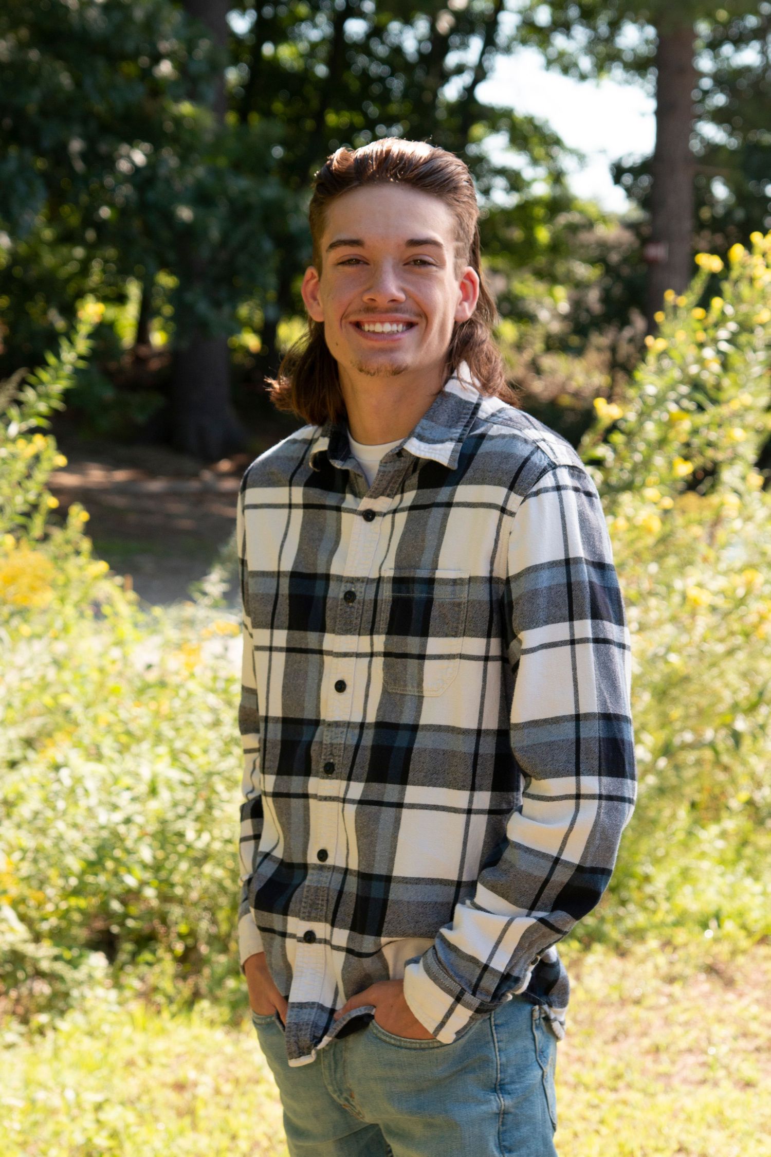 Smiling young man with mullet hairstyle wearing plaid flannel shirt standing outdoors in sunny garden setting.
