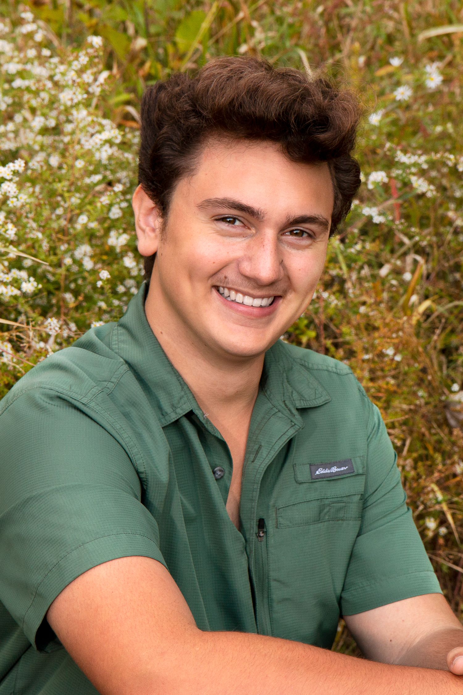 Smiling young man in green Eddie Bauer shirt posing outdoors among wildflowers for senior portrait.