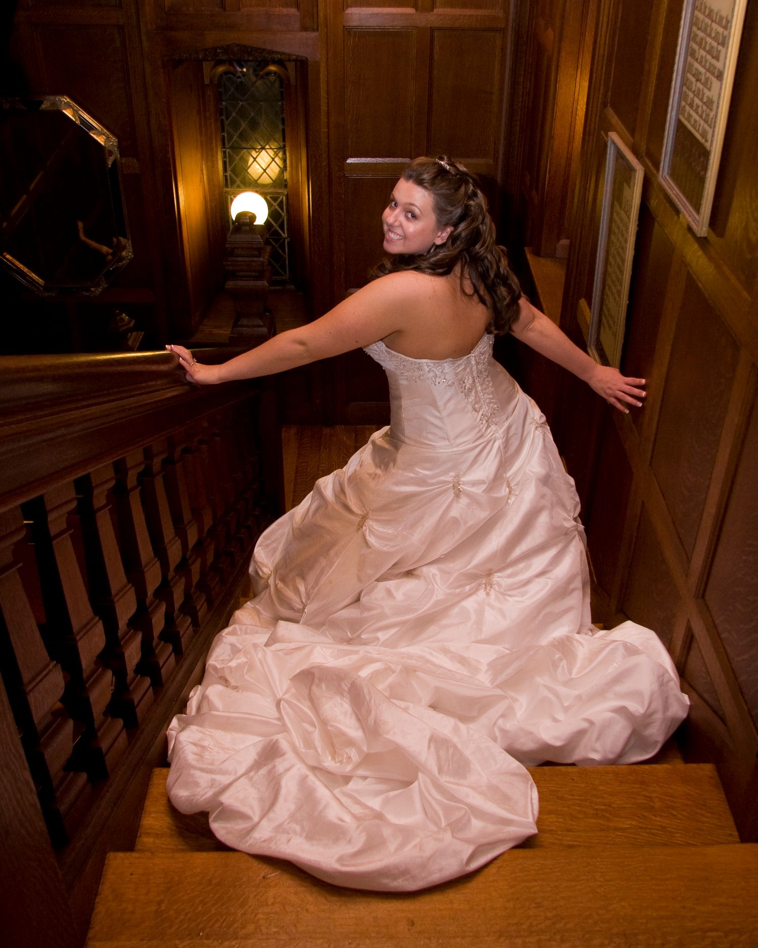 Bride in strapless ballgown descends elegant wooden staircase in warmly lit interior.