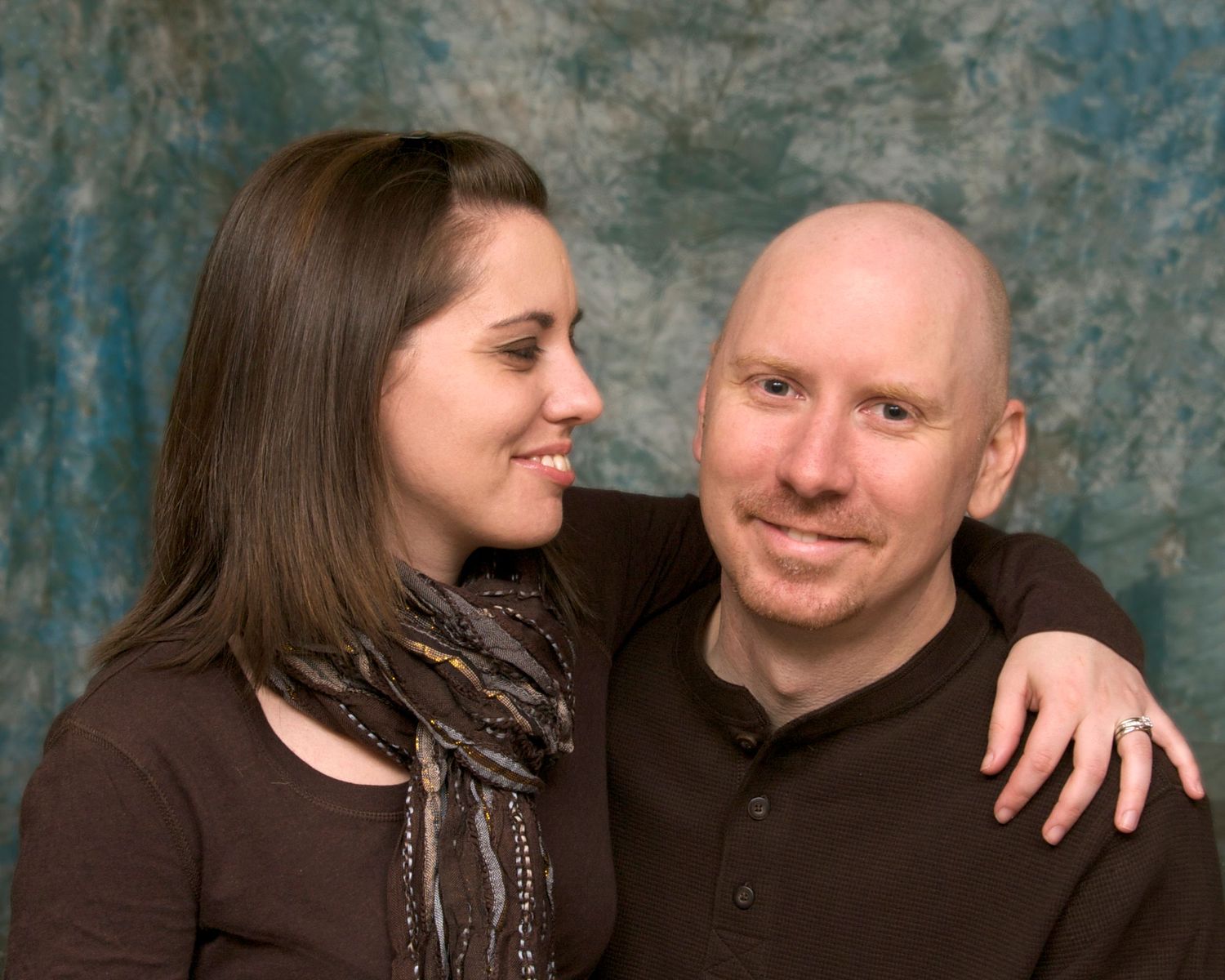 A smiling couple poses together against a blue-gray backdrop, the woman resting her arm around the bald mans shoulder.