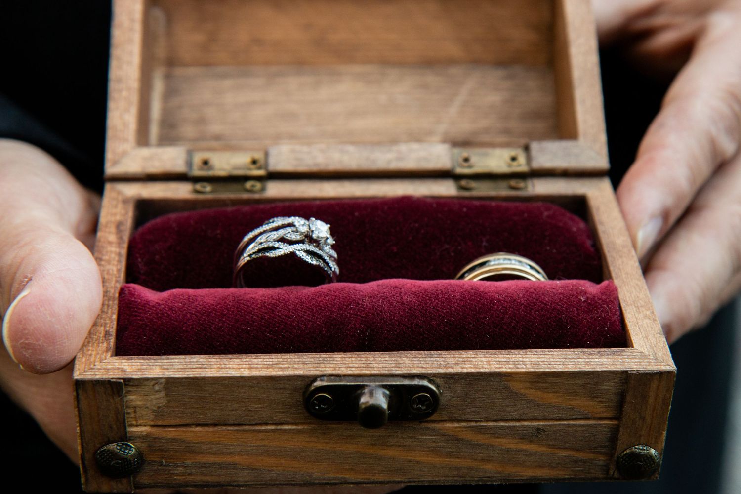 Hands holding rustic wooden box with red velvet interior displaying diamond wedding rings.