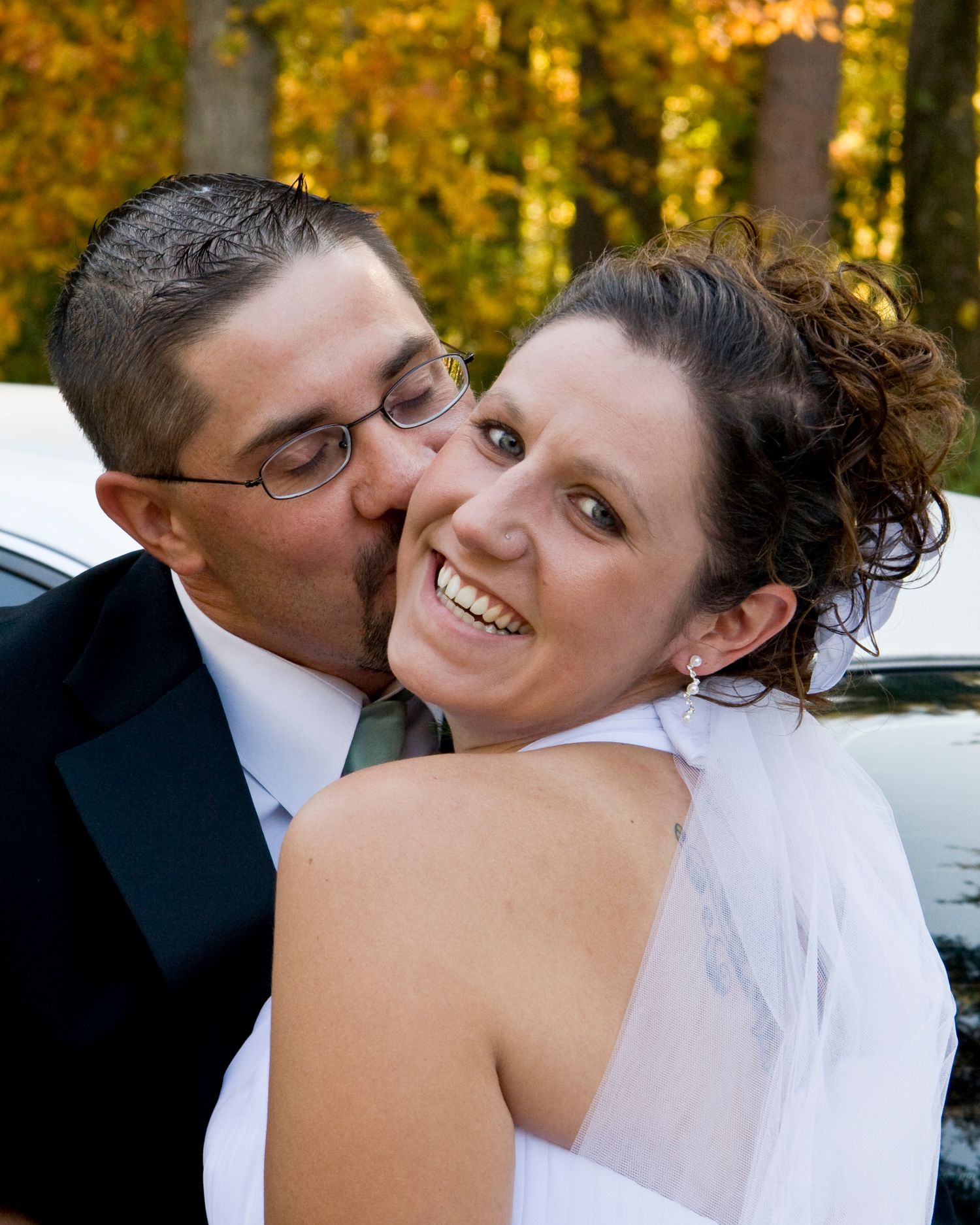 Groom kisses smiling bride on cheek during outdoor fall wedding photo session.
