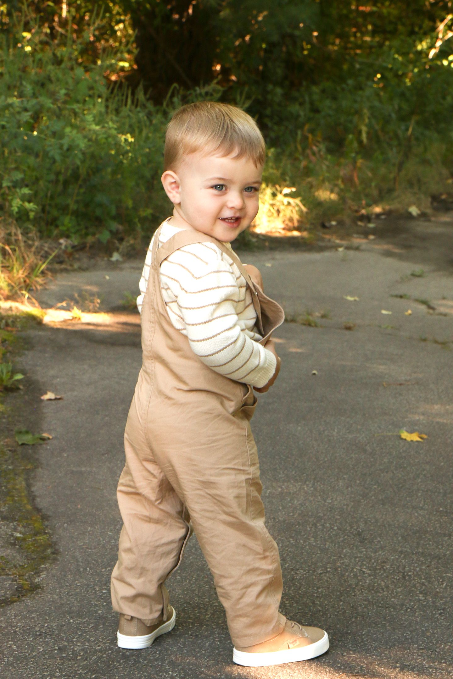 Happy toddler boy in tan overalls and striped shirt smiling while walking on outdoor path.