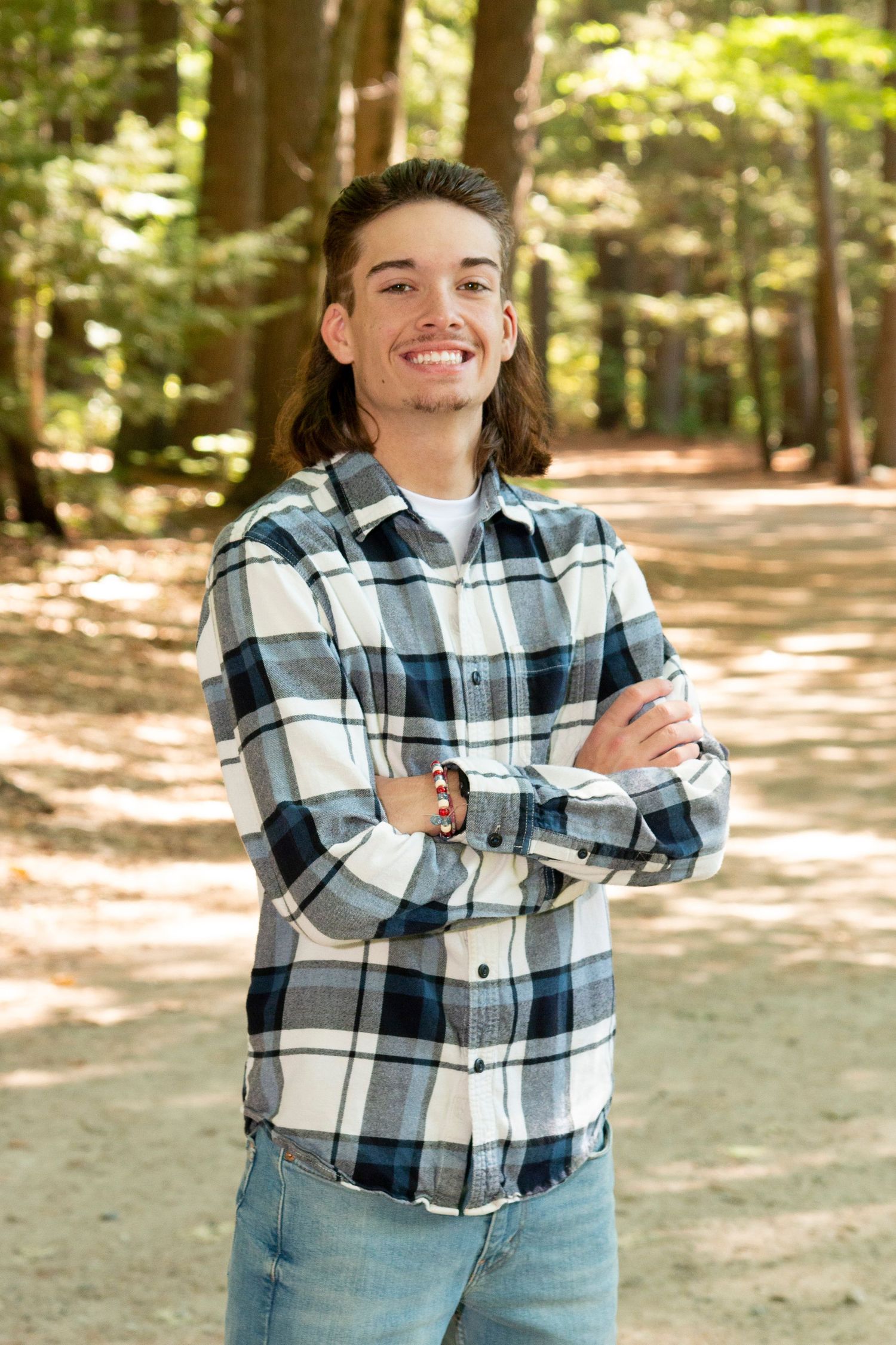 Smiling young man with long hair in plaid shirt standing with arms crossed in forest.