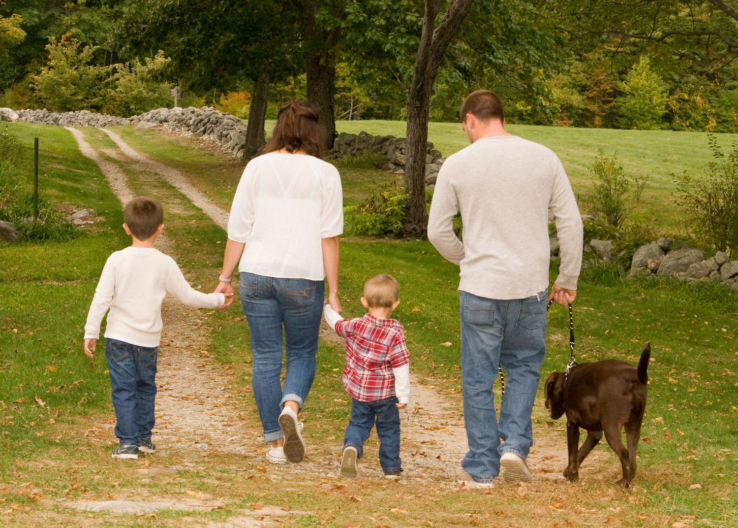 Family of four with dog walks hand-in-hand down a scenic gravel path surrounded by autumn trees.