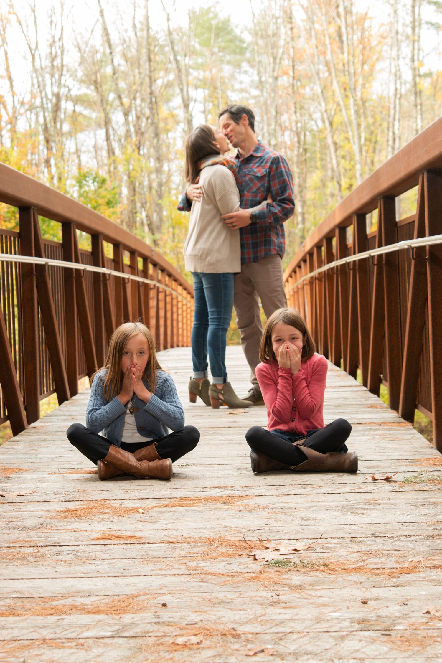 Two girls covering mouths while parents kiss on rustic wooden bridge surrounded by autumn foliage.