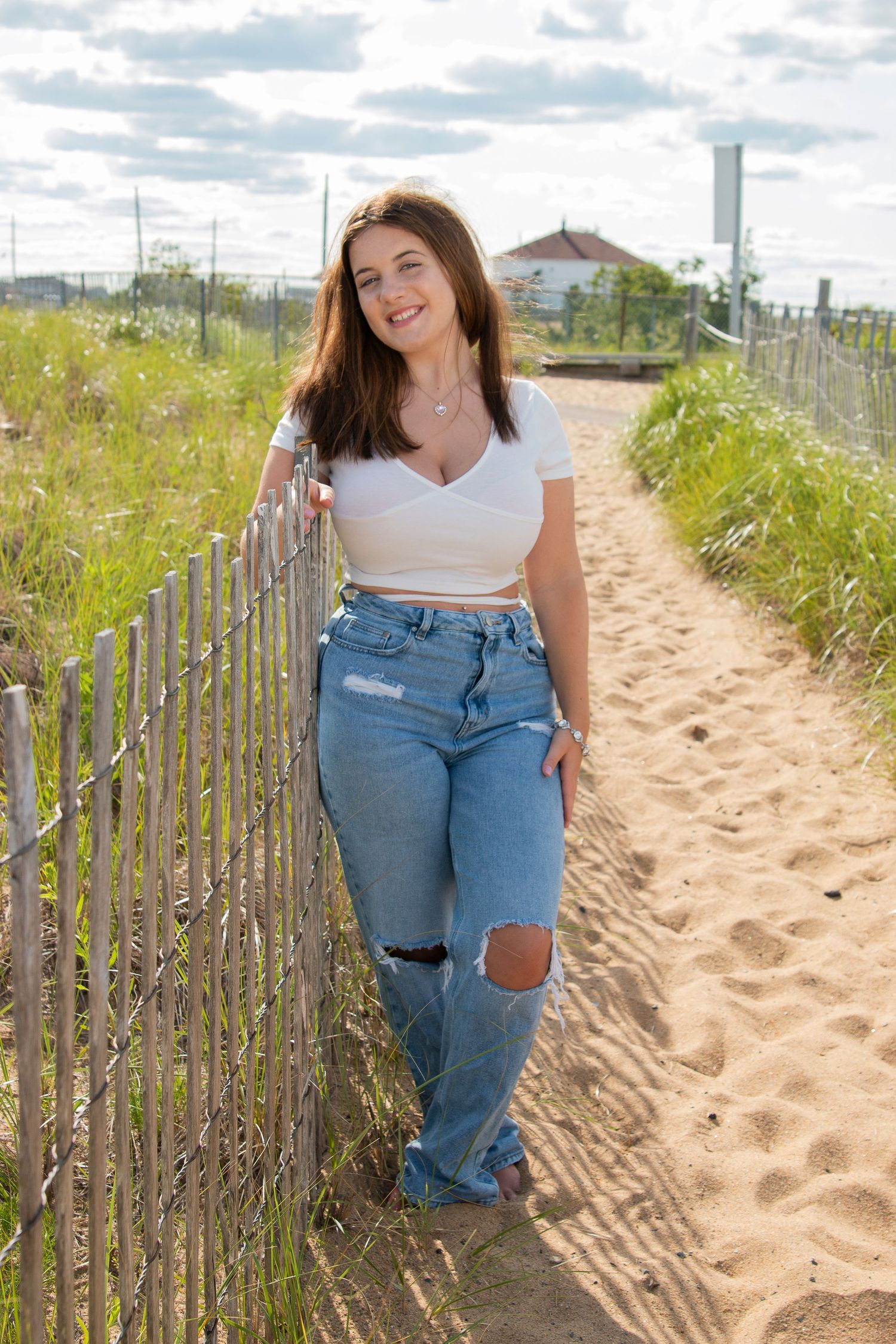 Young woman in white crop top and ripped jeans leaning on wooden fence along sandy beach pathway.