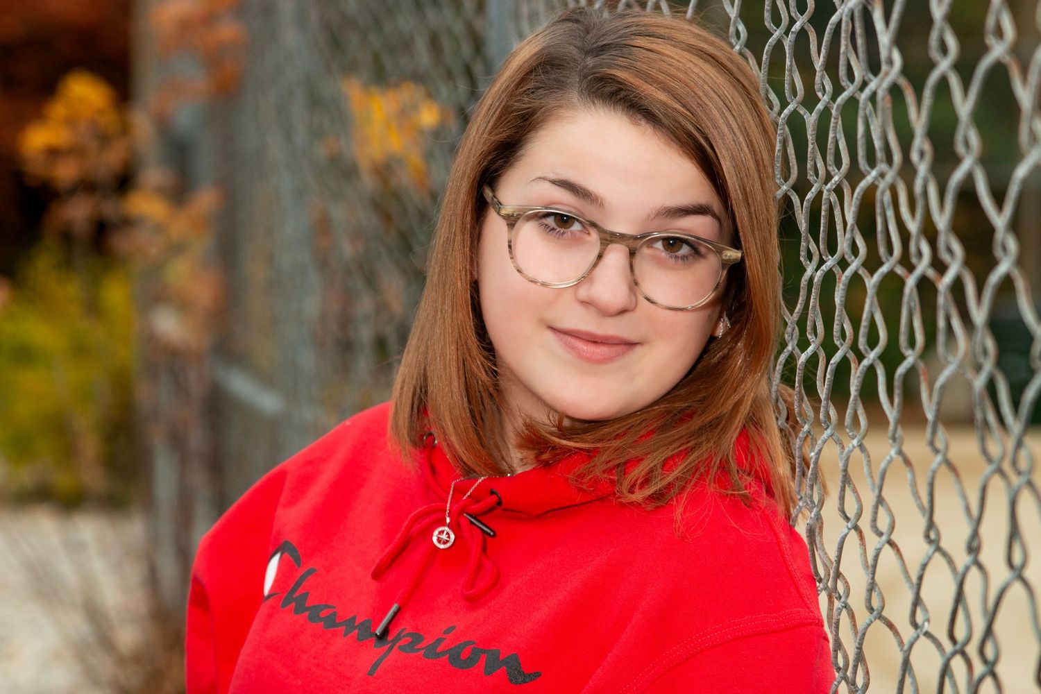 Young teenage girl wearing glasses and red Champion hoodie leaning against chain-link fence.