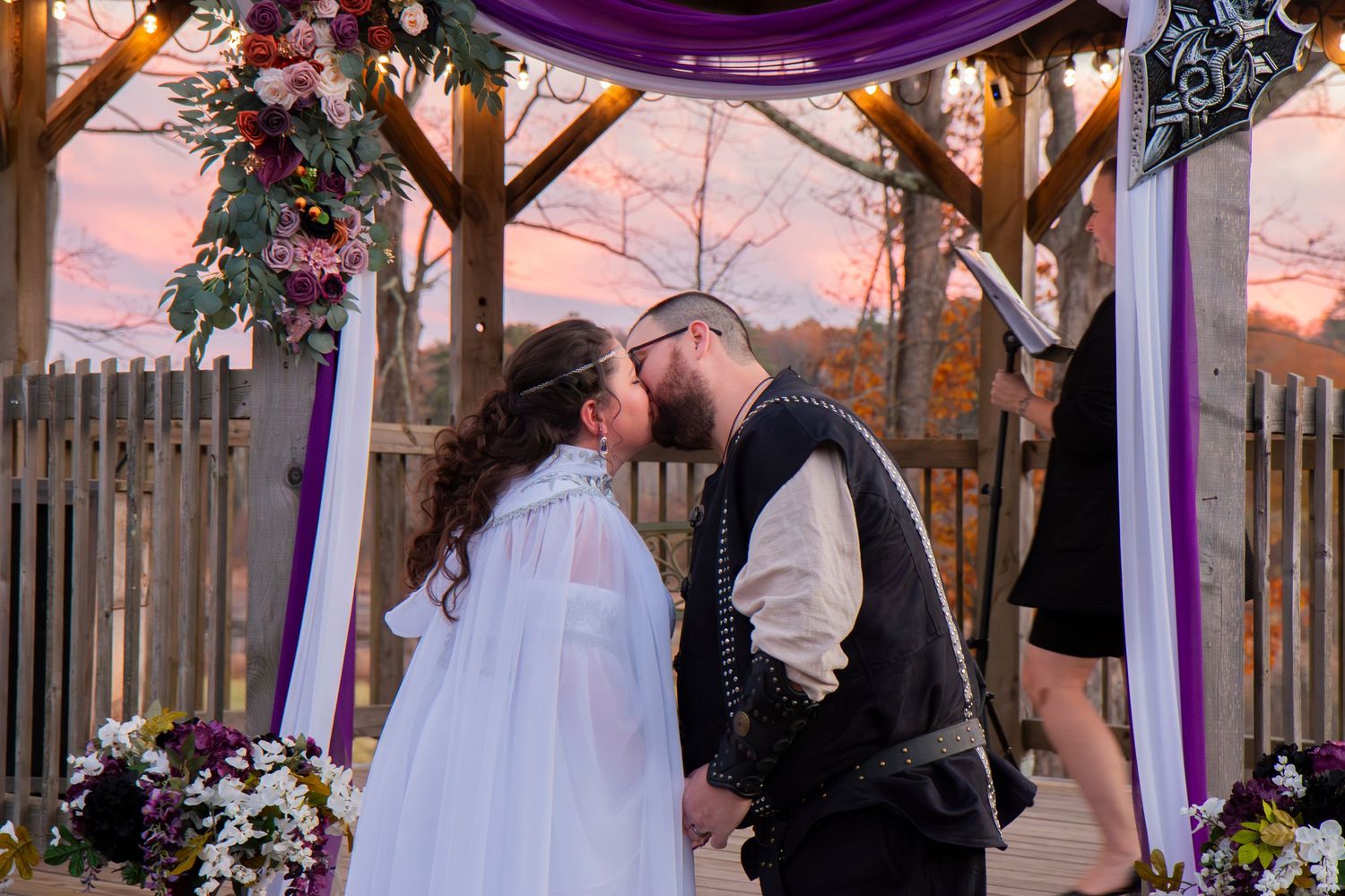 Couple sharing first kiss under floral wedding arch at sunset with purple decorations.