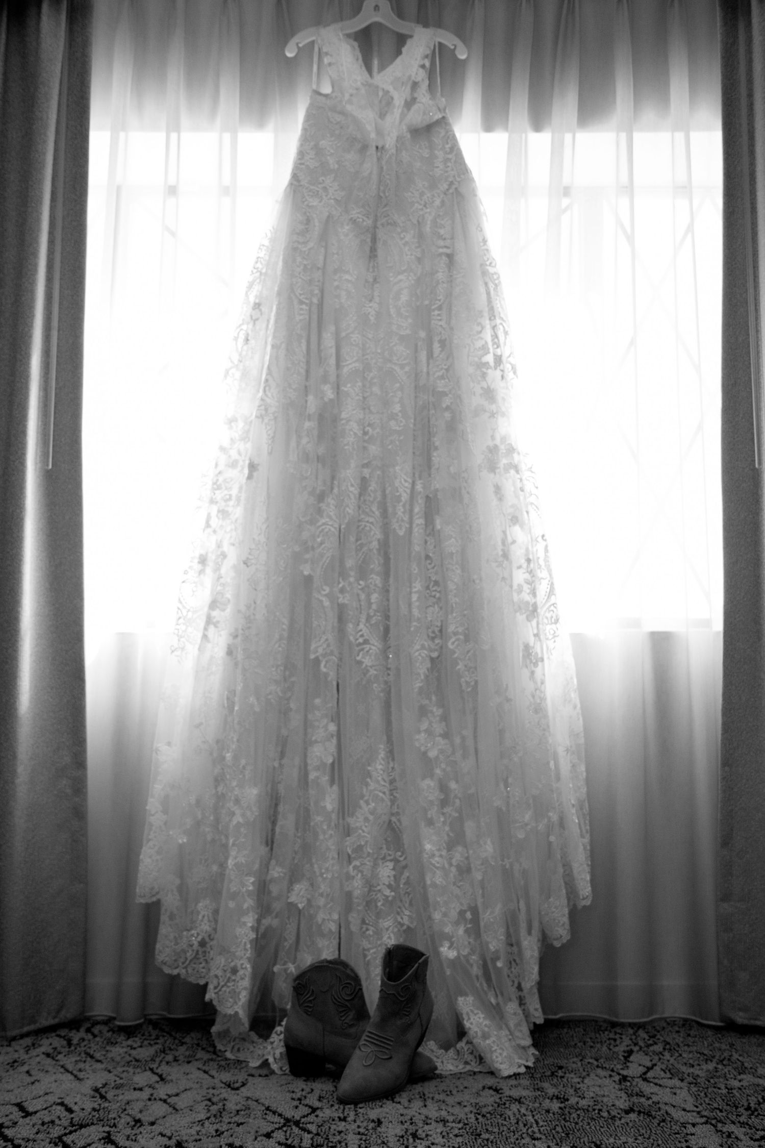 Elegant lace wedding gown hanging in window with cowboy boots below in black and white photo.