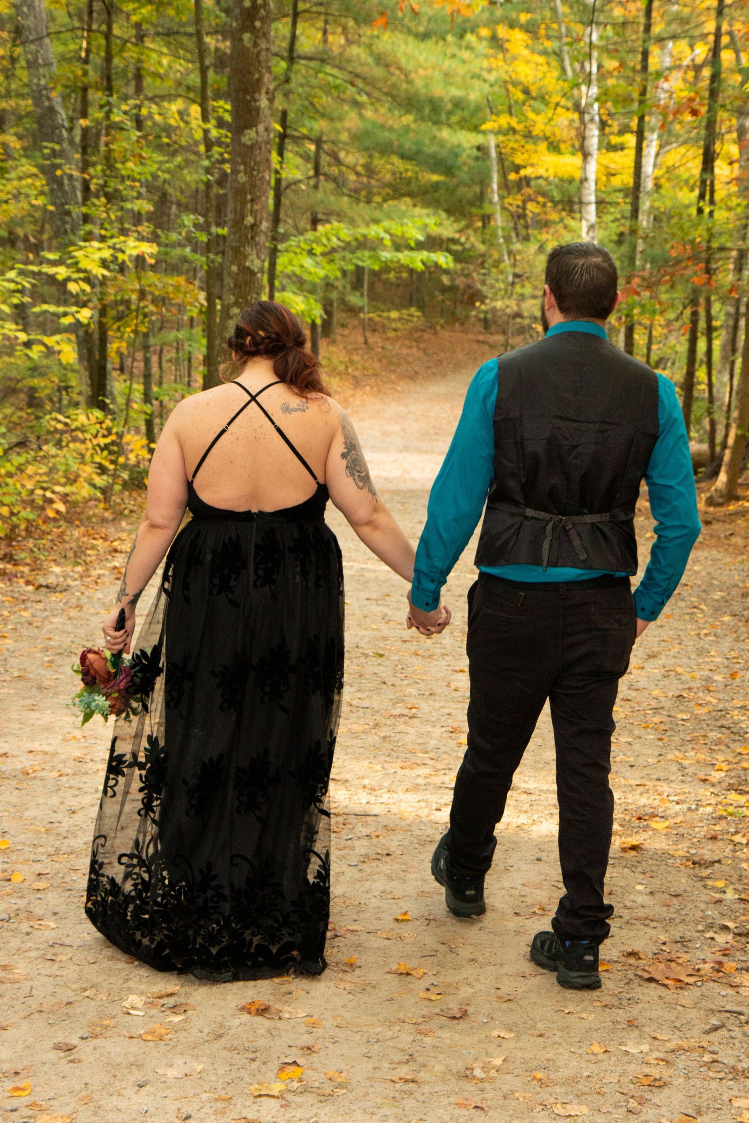 Couple holding hands walking on autumn forest path, bride in black backless gown with bouquet, groom in teal shirt and vest.