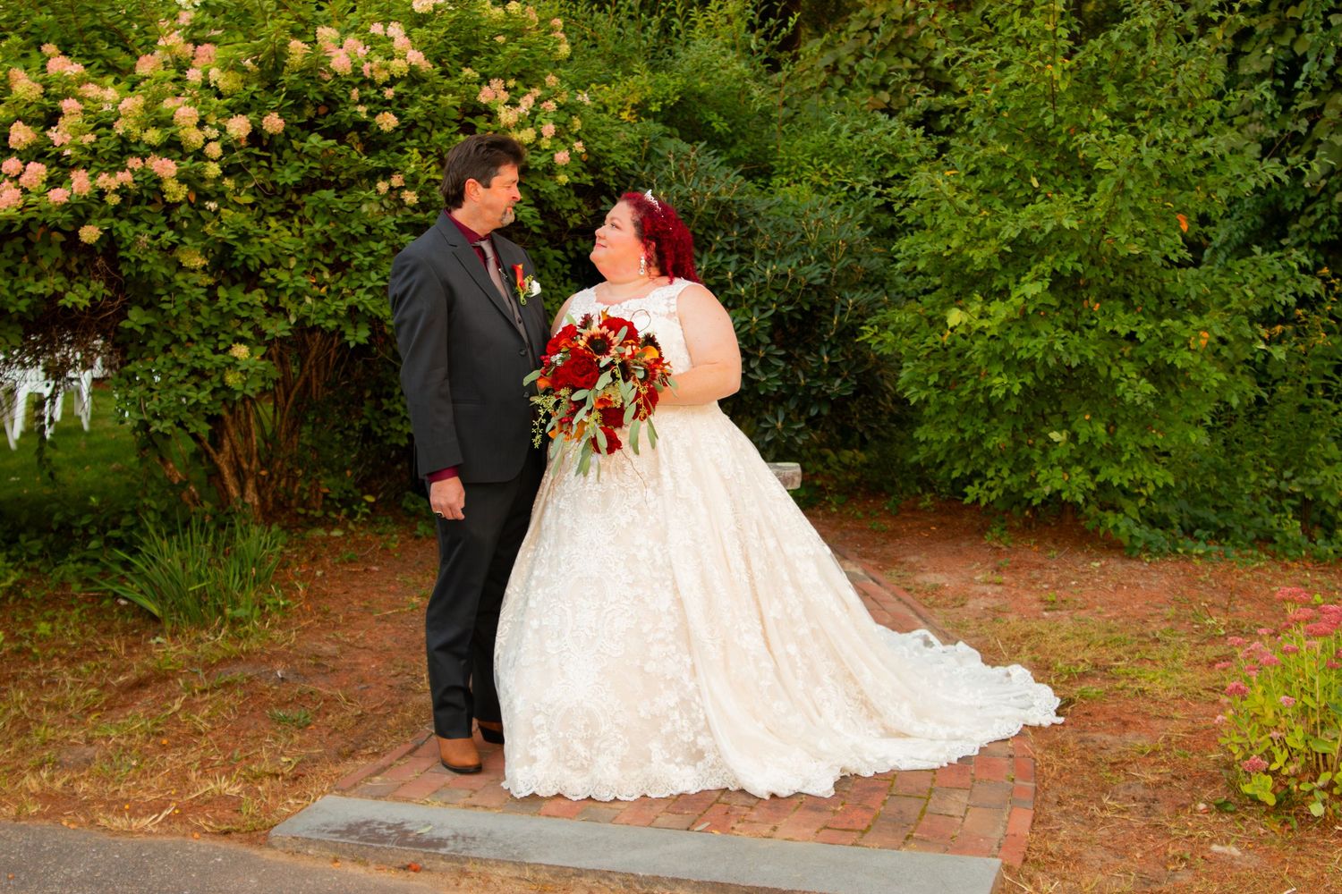 Bride in lace ball gown with red bouquet smiles at groom outdoors near blooming shrubs.