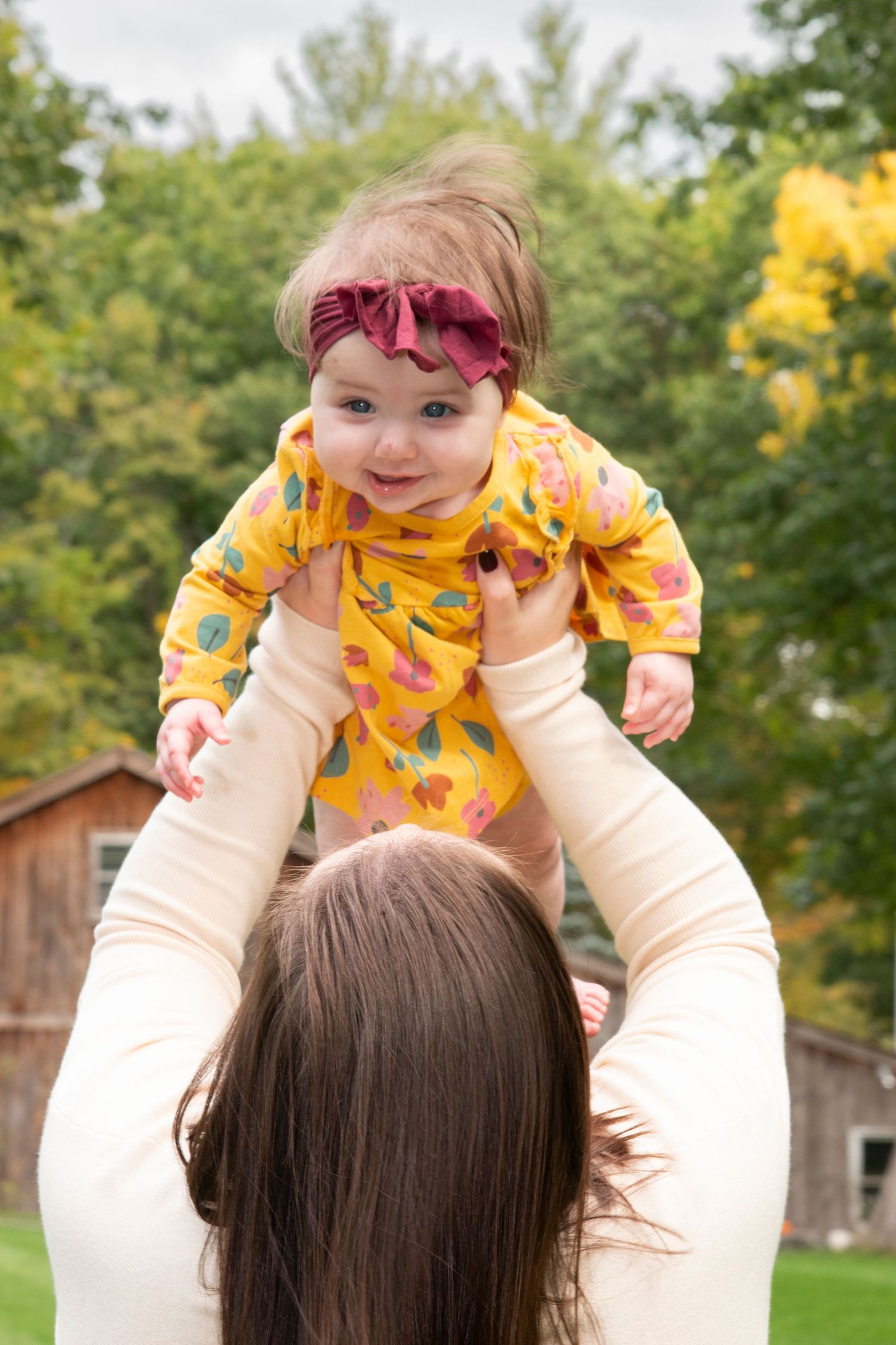 A smiling baby in a yellow floral outfit with a pink bow headband being lifted up by her mother outdoors.