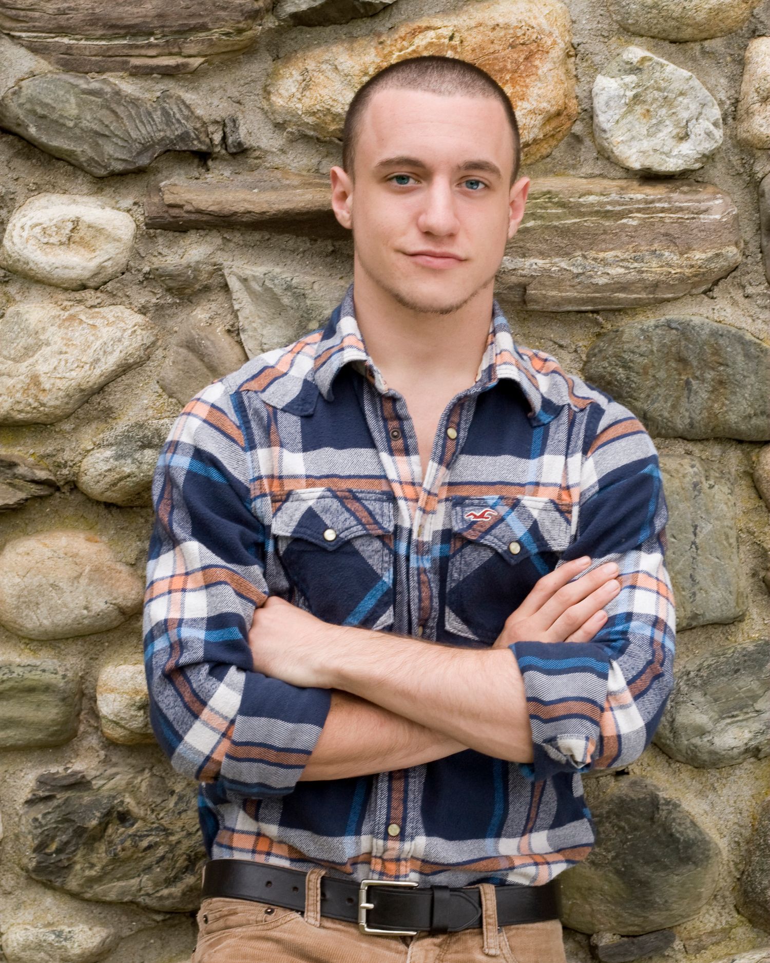 Young man in plaid flannel shirt stands arms crossed against stone wall outdoors.