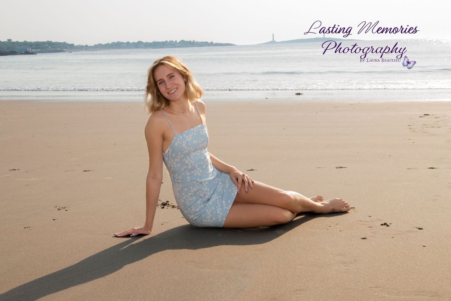 Blonde teenage girl in blue dress sitting on sandy beach for senior portrait photography.