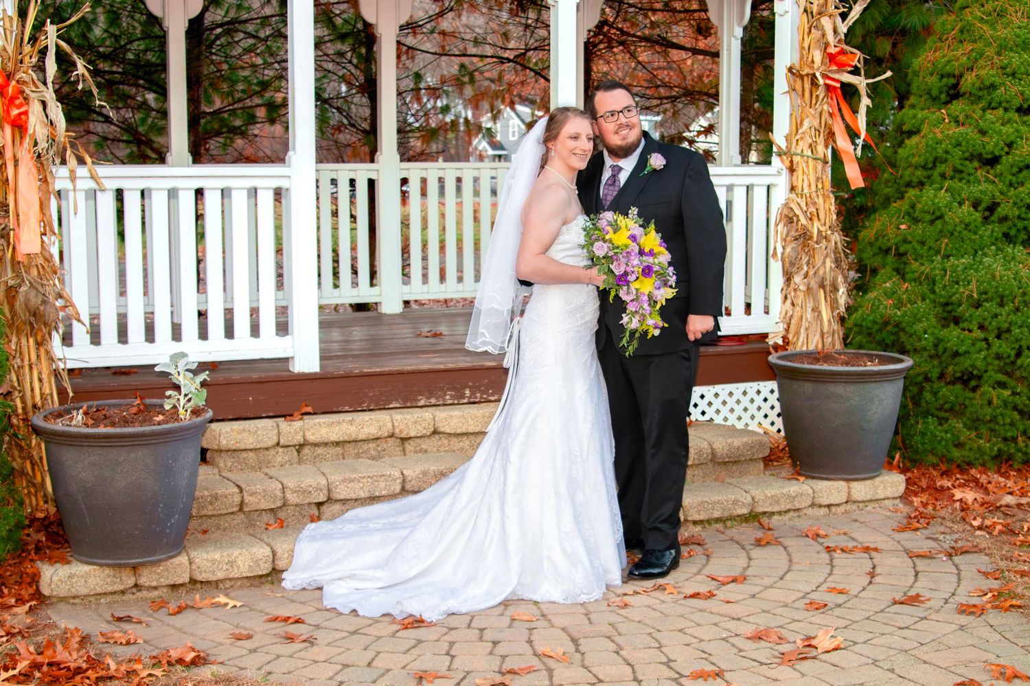 Bride and groom posing in front of a white gazebo decorated with fall corn stalks on a brick patio.
