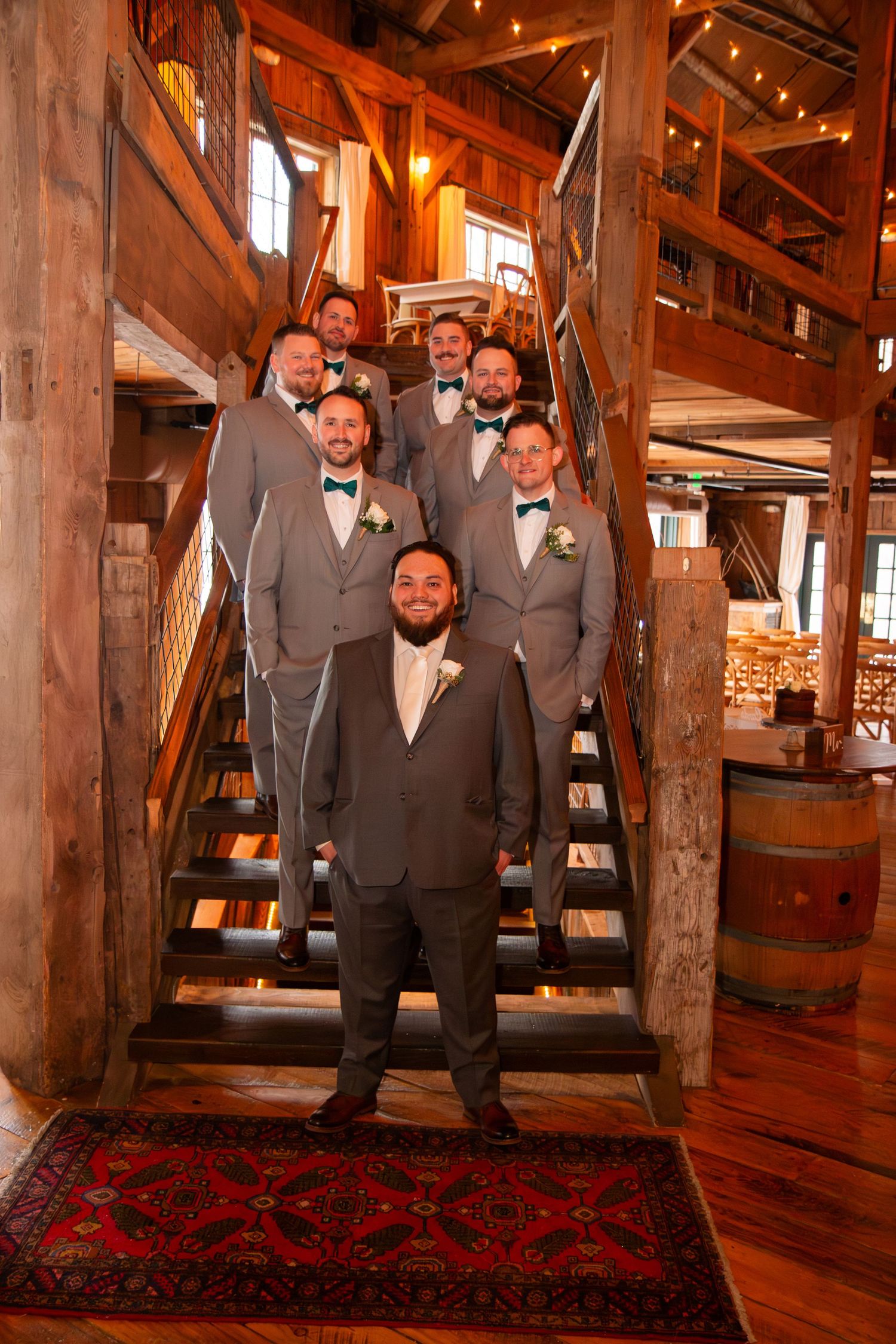 Groom and groomsmen in gray suits with teal bow ties pose on rustic barn staircase.