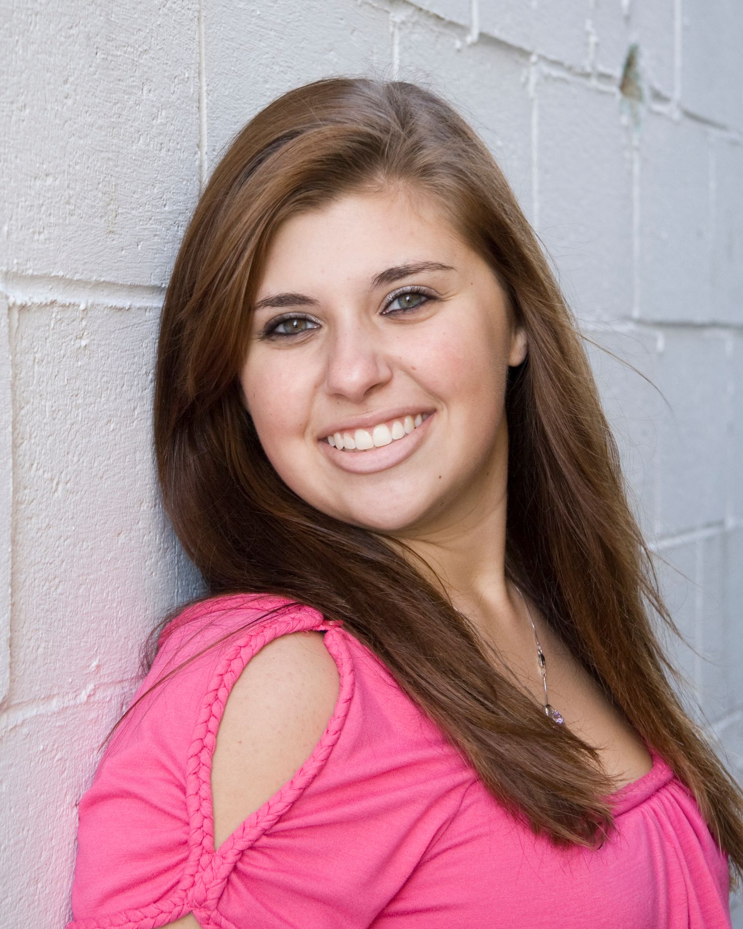 Smiling brunette teen girl in pink top leans against white concrete block wall.