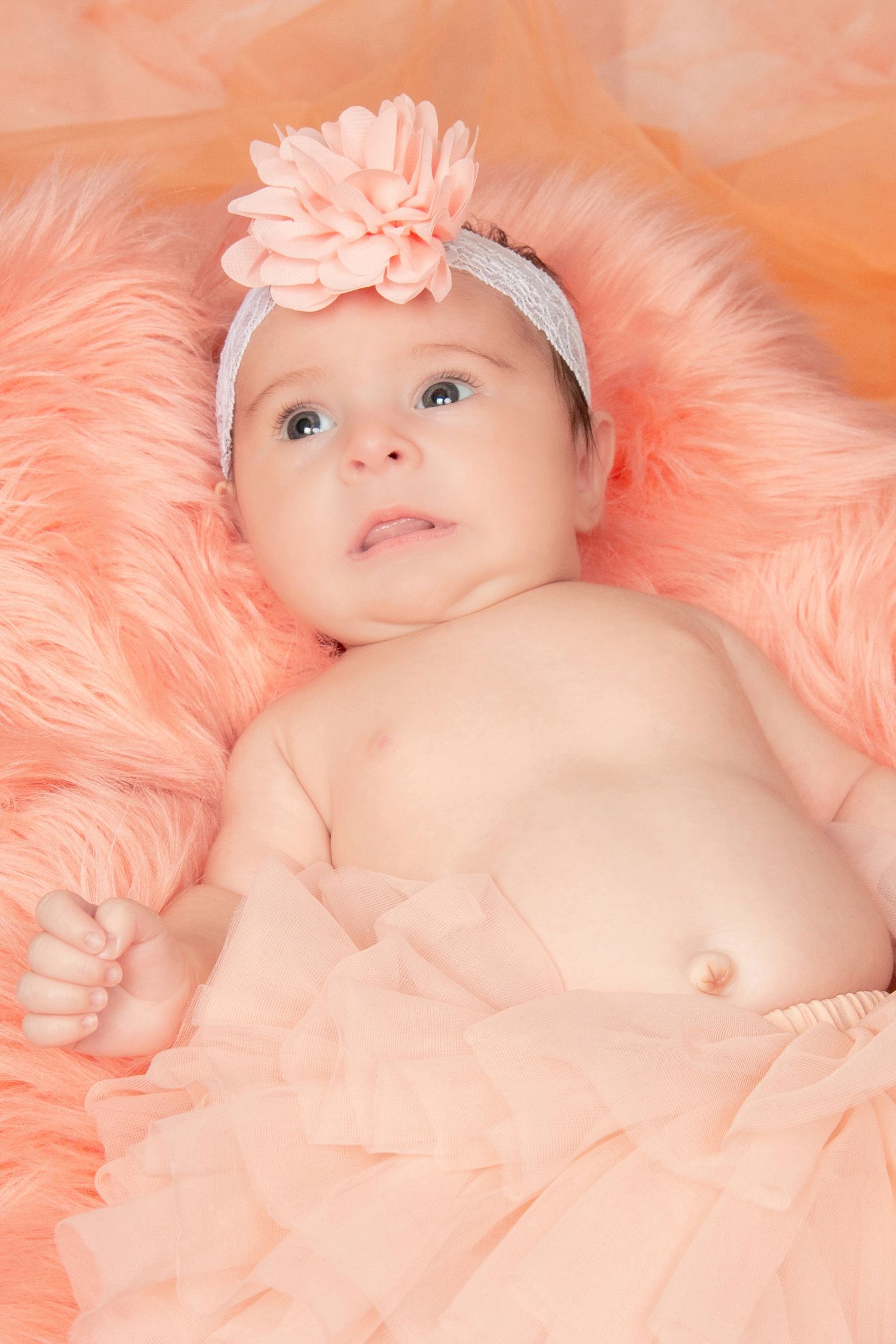 Sweet baby girl in peach tutu with flower headband lying on fluffy pink fur blanket.