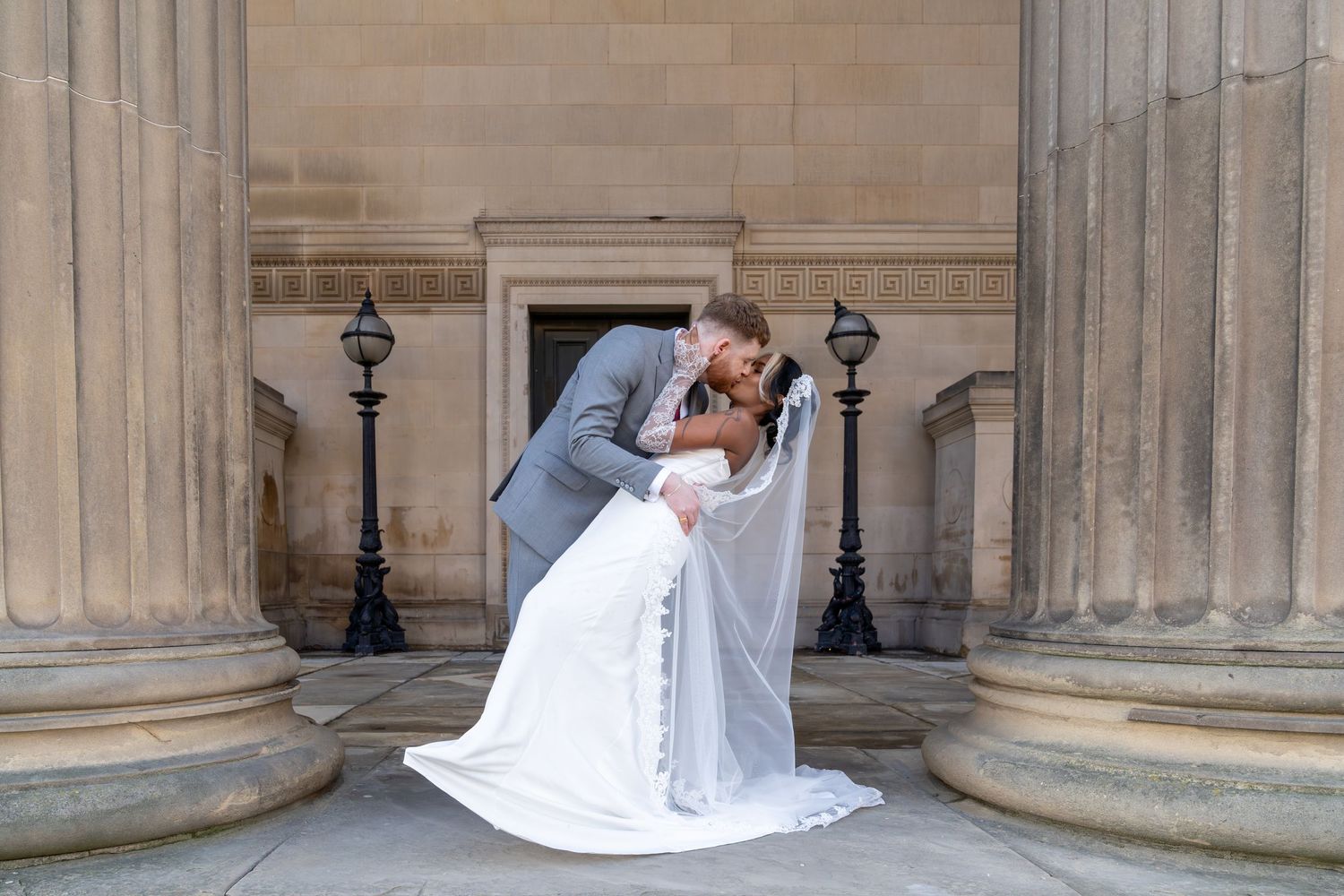 A romantic wedding couple shares a tender moment between classic stone columns at a grand historic building entrance.