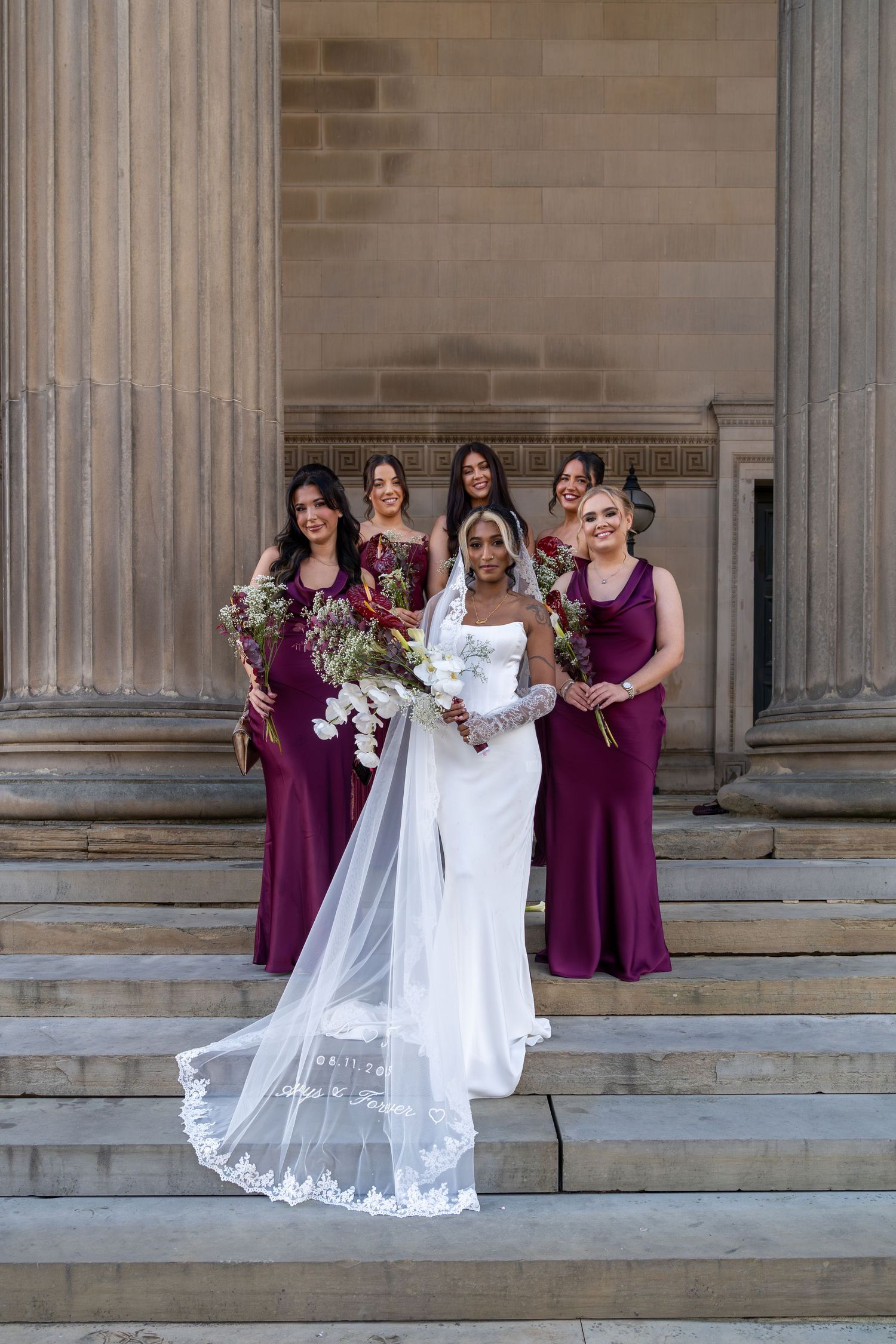 Wedding party poses on stone steps with bride in white gown and bridesmaids in burgundy dresses at elegant venue.