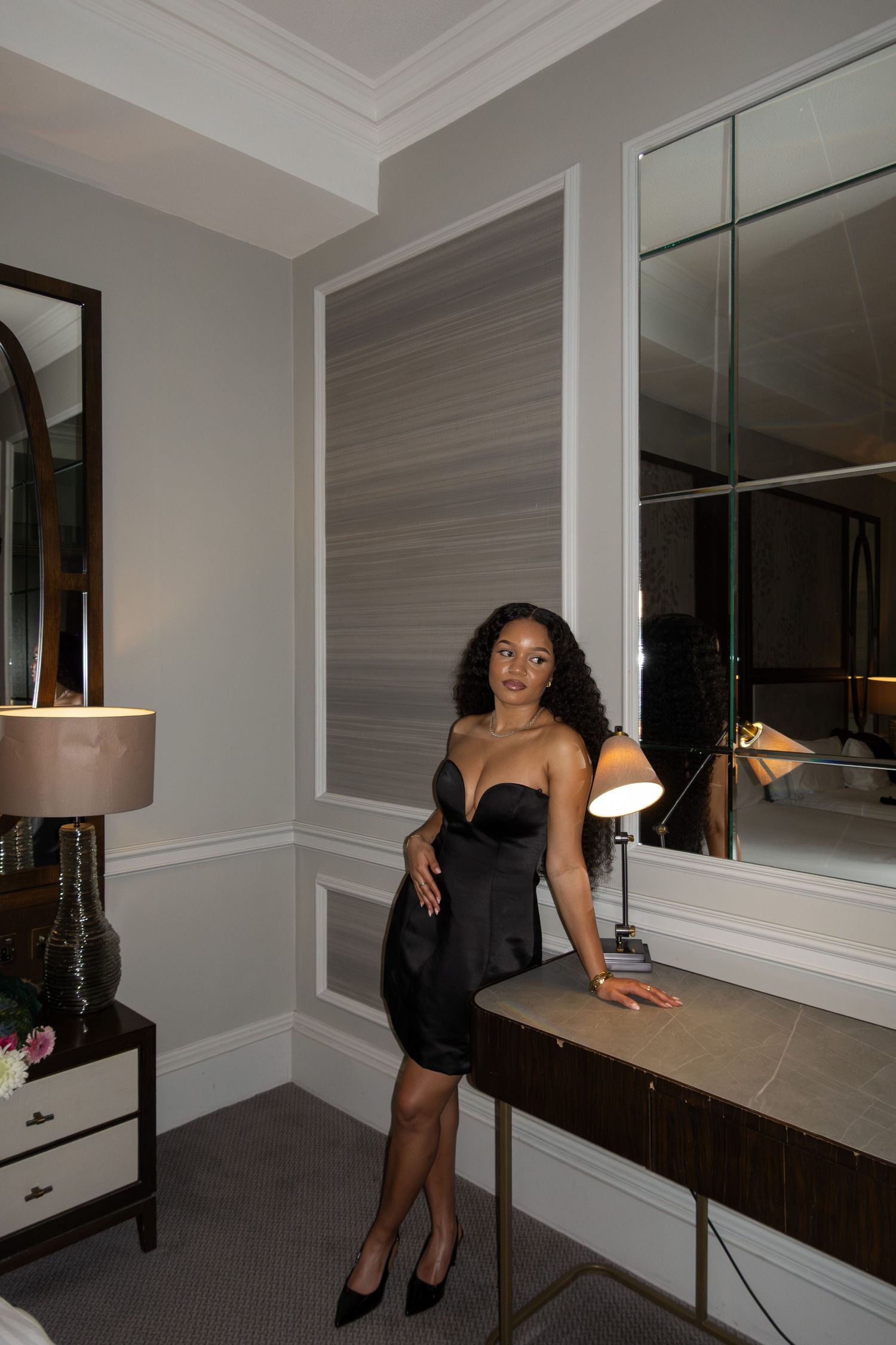 Woman in elegant black strapless dress posing in a luxury hotel room with mirror and lamp.