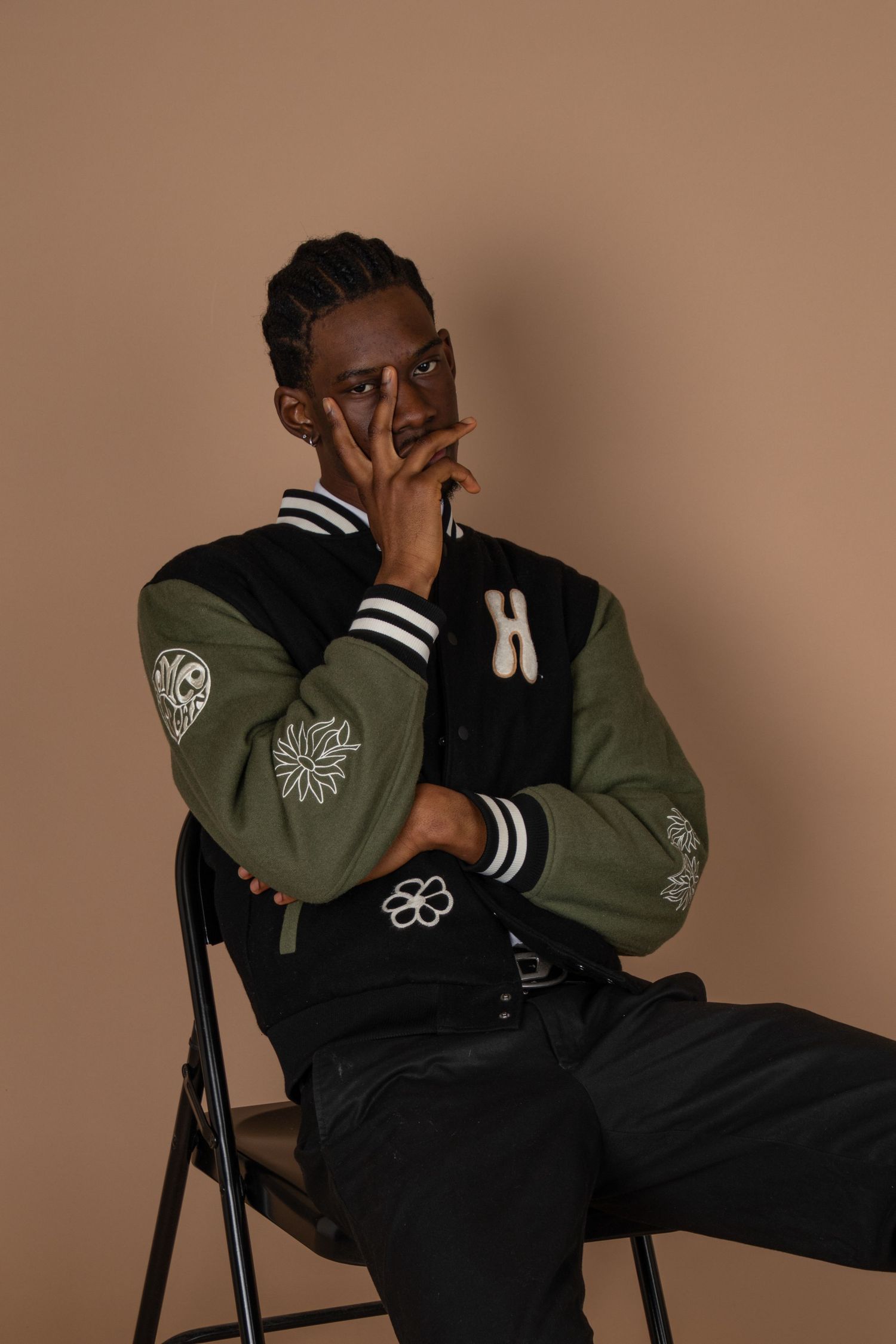 Person in olive varsity jacket sitting on chair against brown backdrop.