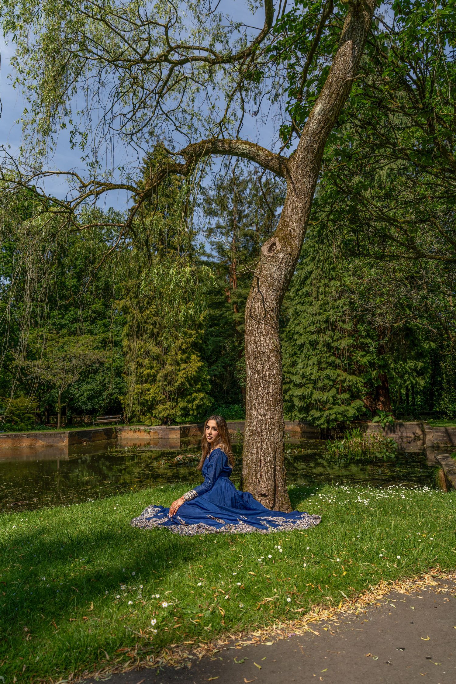 Woman in blue dress sitting against a tree by a pond in a lush green park on a sunny day.