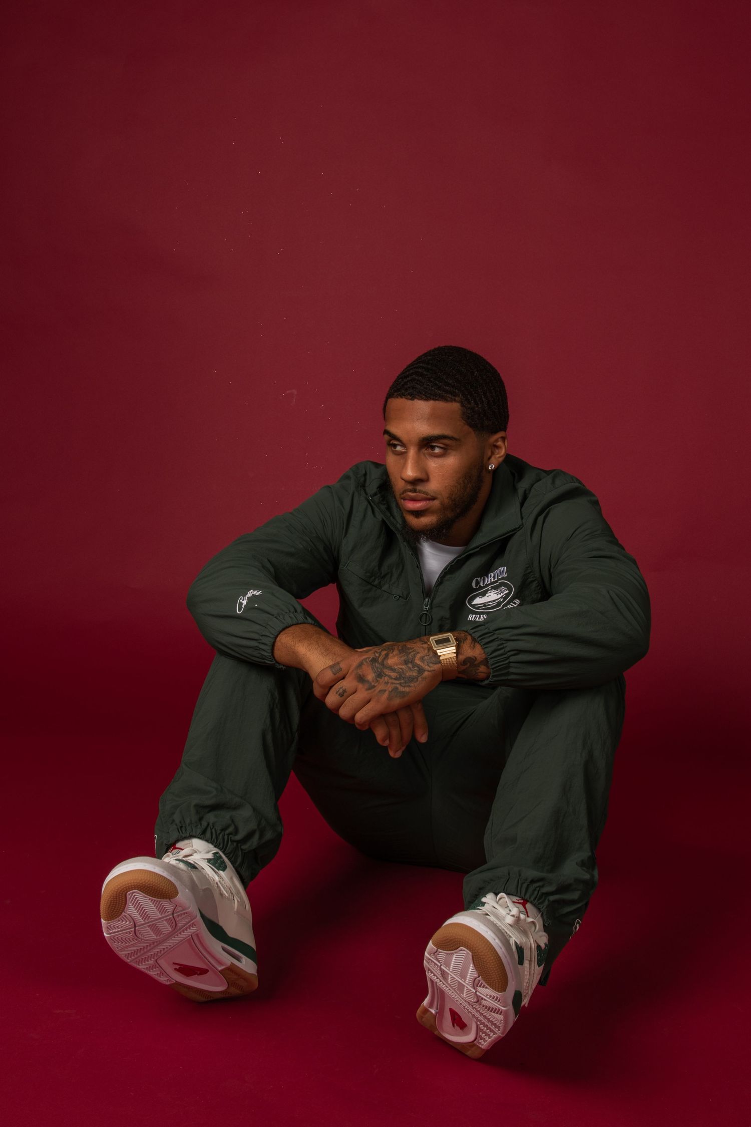 Athletic figure in dark clothing sitting cross-legged against burgundy backdrop.