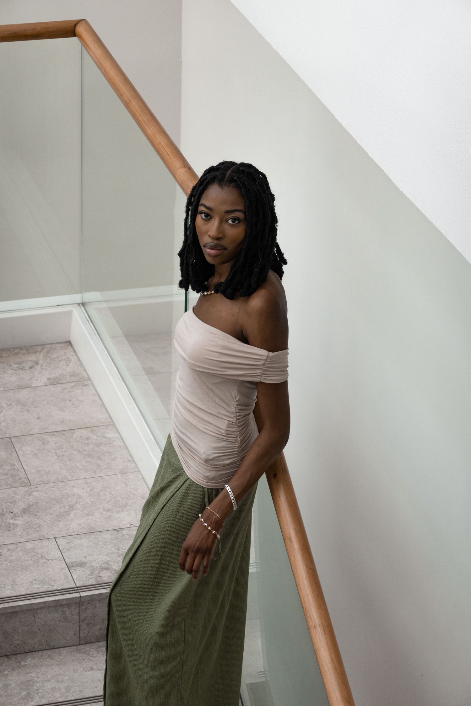 Fashion model in off-shoulder top and olive skirt poses elegantly on modern staircase.