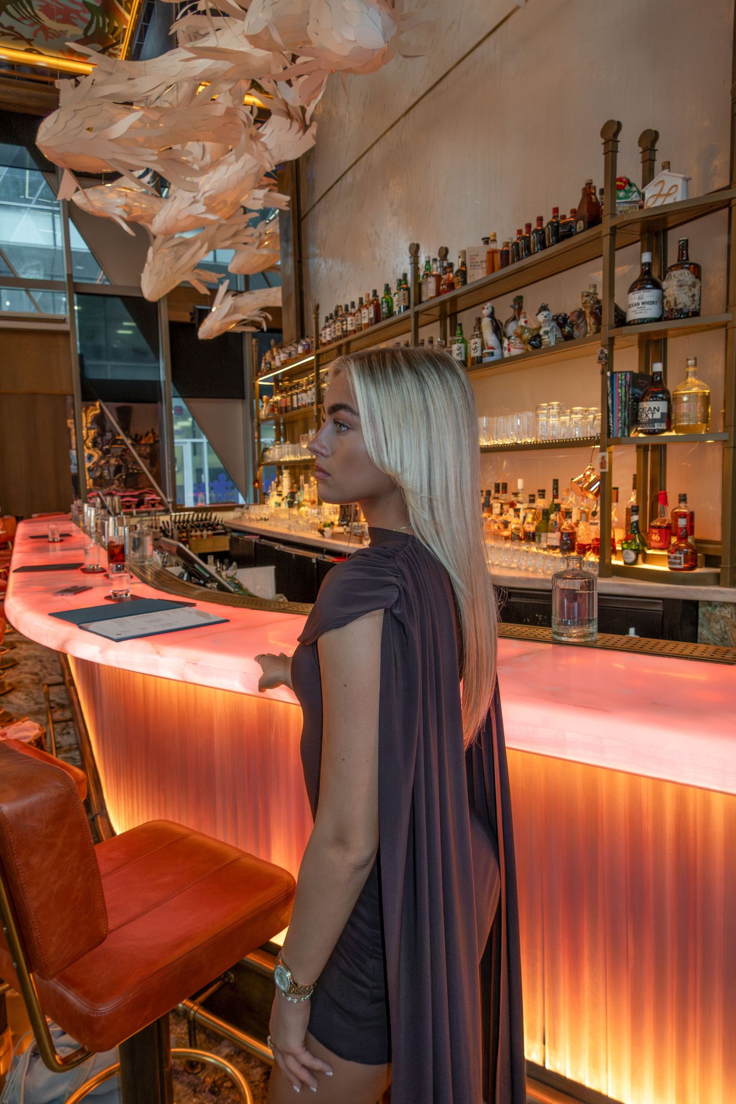 Blonde woman in backless gray gown stands at illuminated pink bar with bottles on shelves behind her.
