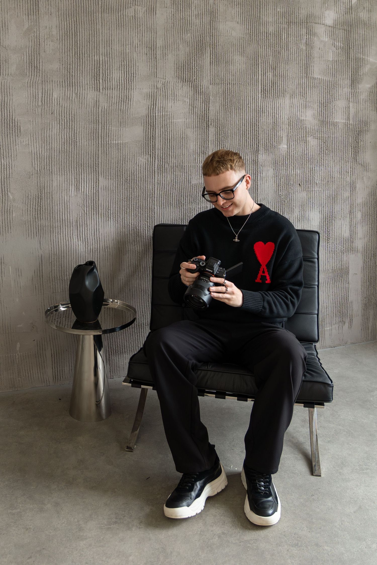 Person in black outfit examining camera while seated in minimalist setting.