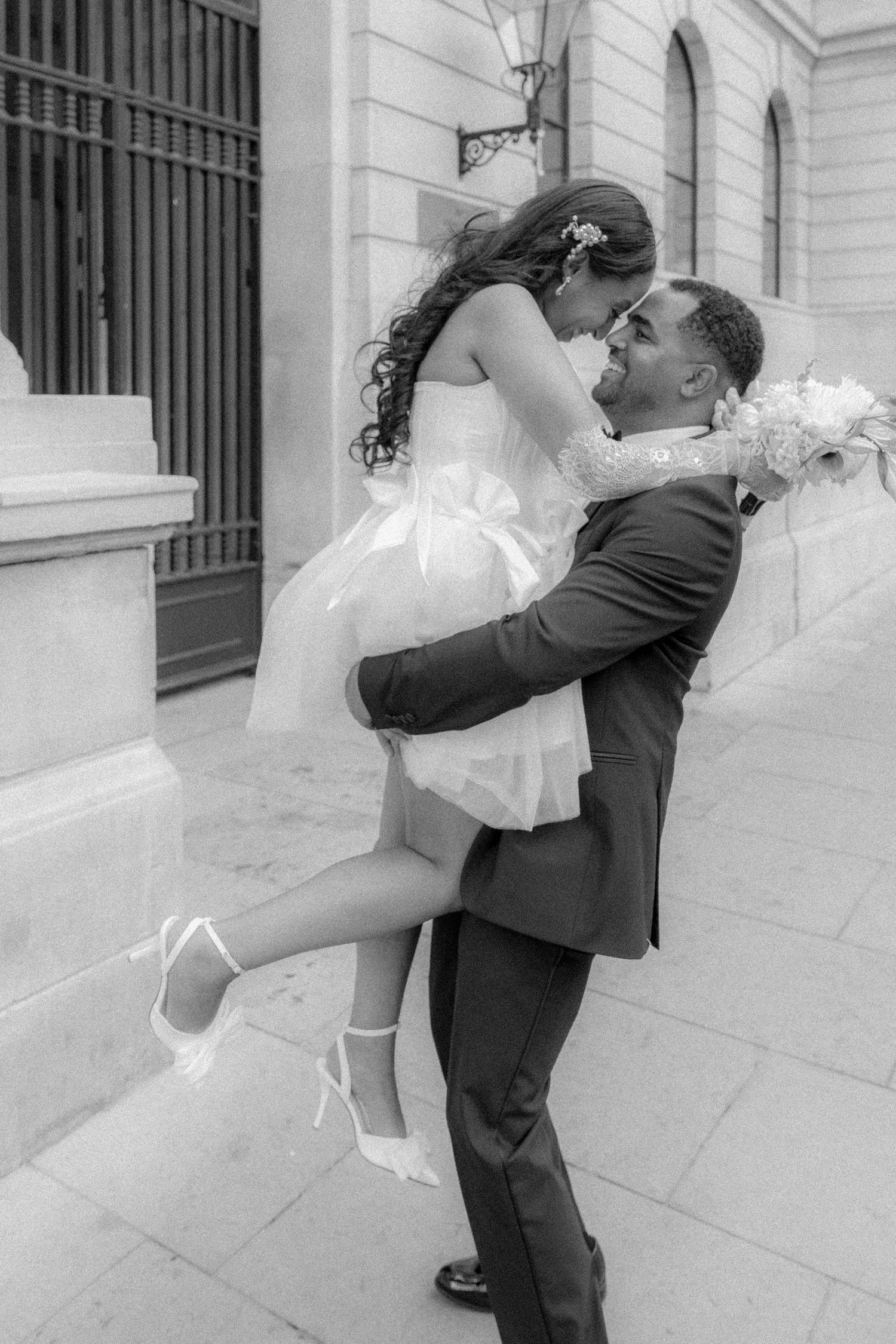 Black and white photo of groom lifting bride in short wedding dress, sharing romantic moment on city street.