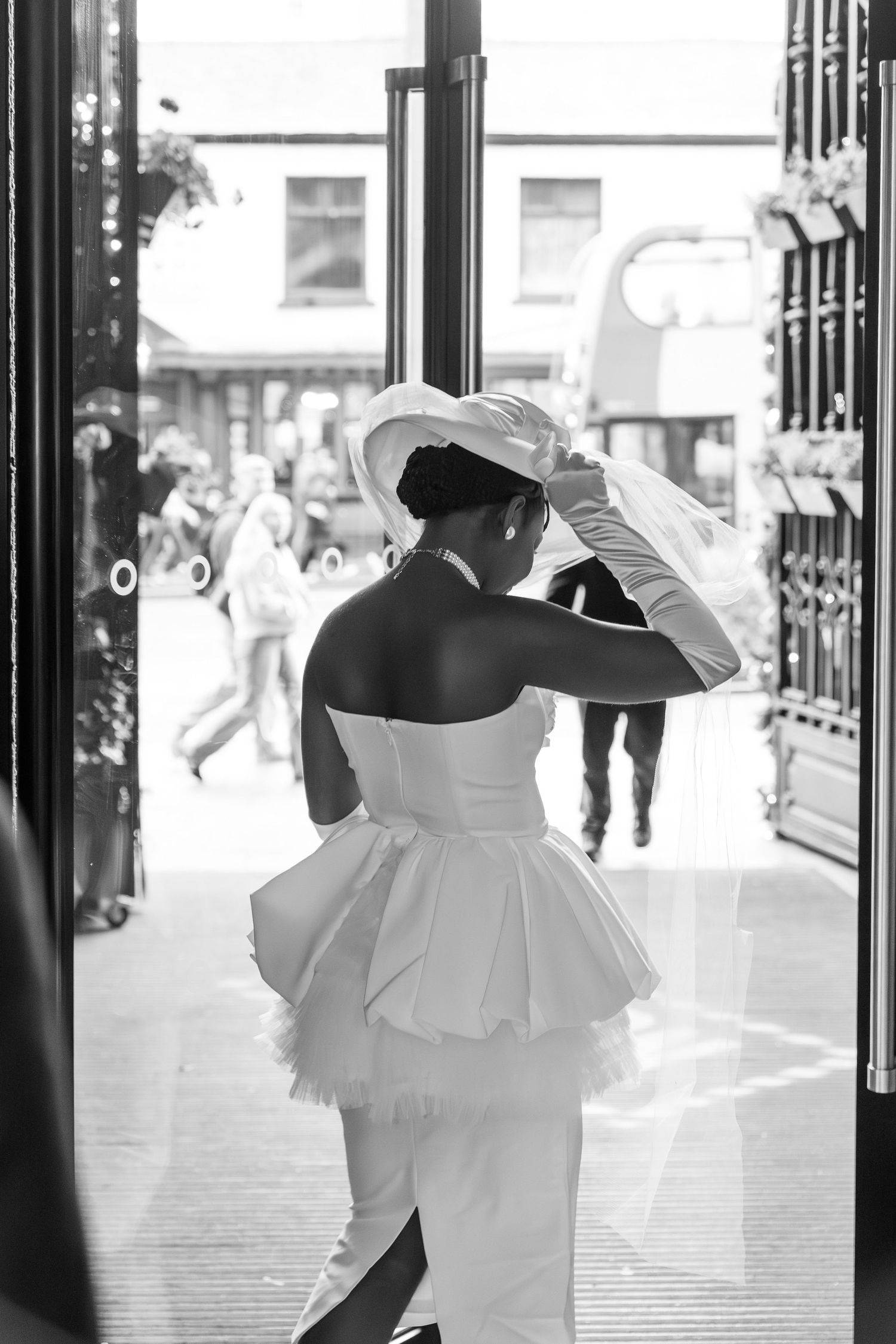 Elegant bride in white strapless gown and wide-brim hat stands in doorway, black and white photography.
