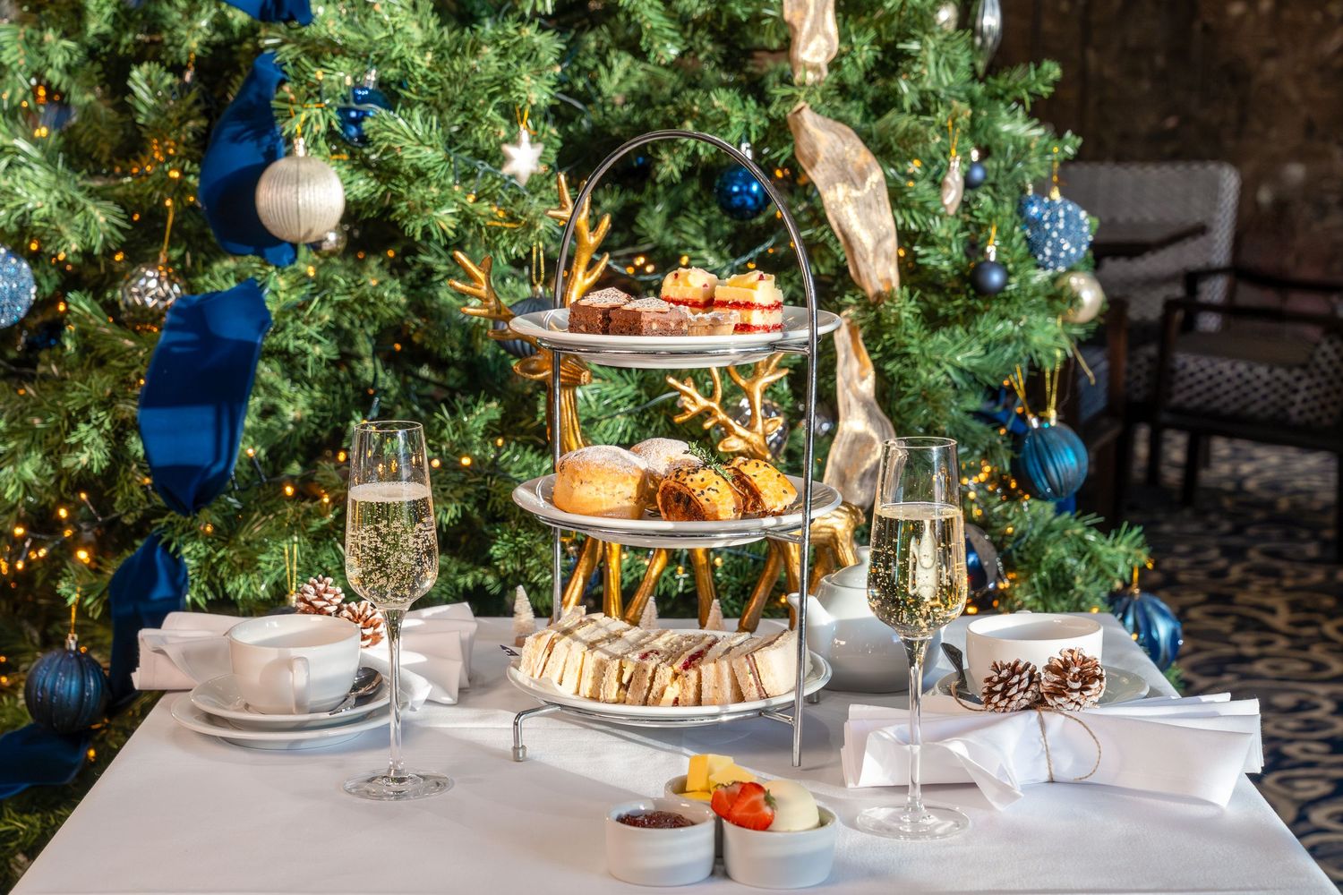 Elegant Christmas table setting with tiered dessert stands, white tablecloth, and evergreen decor beneath twinkling lights.