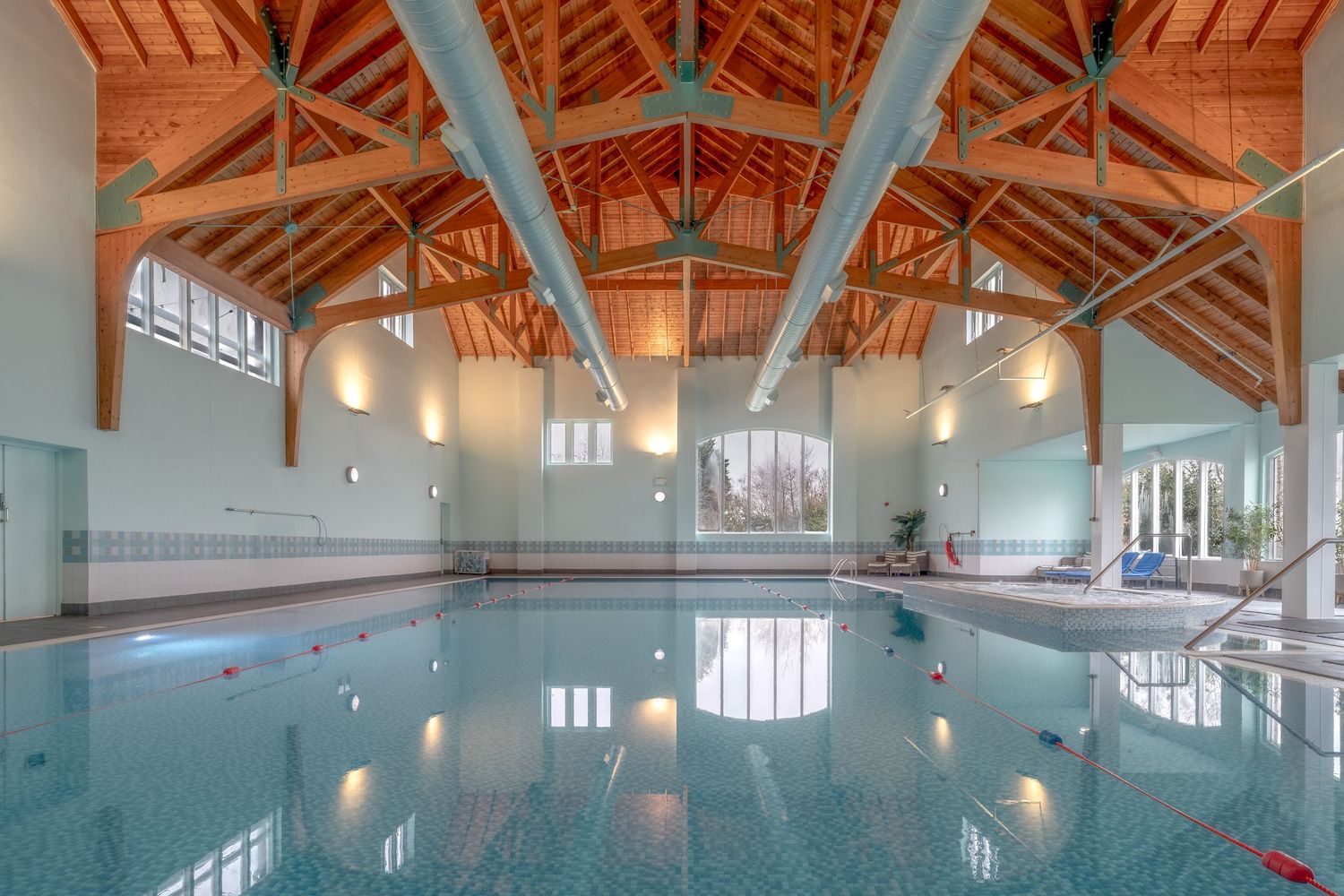 Indoor swimming pool with vaulted wooden beam ceiling, calm reflective water, and large windows in an elegant aquatic facility.