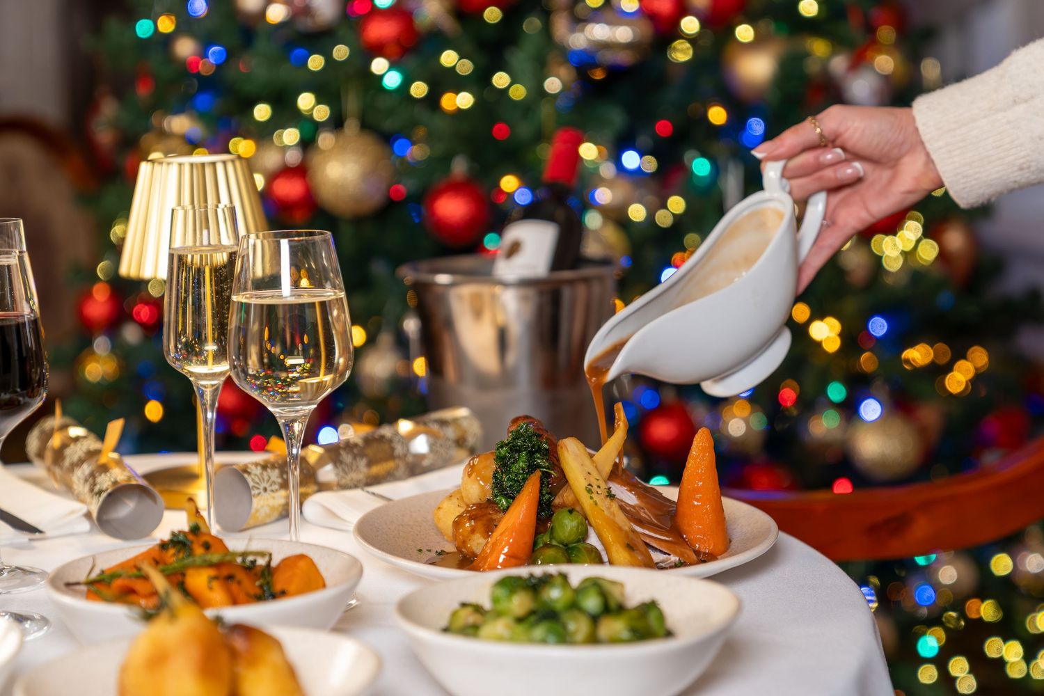Elegant holiday dinner table with colorful Christmas tree lights glowing in the background and plated gourmet meals.