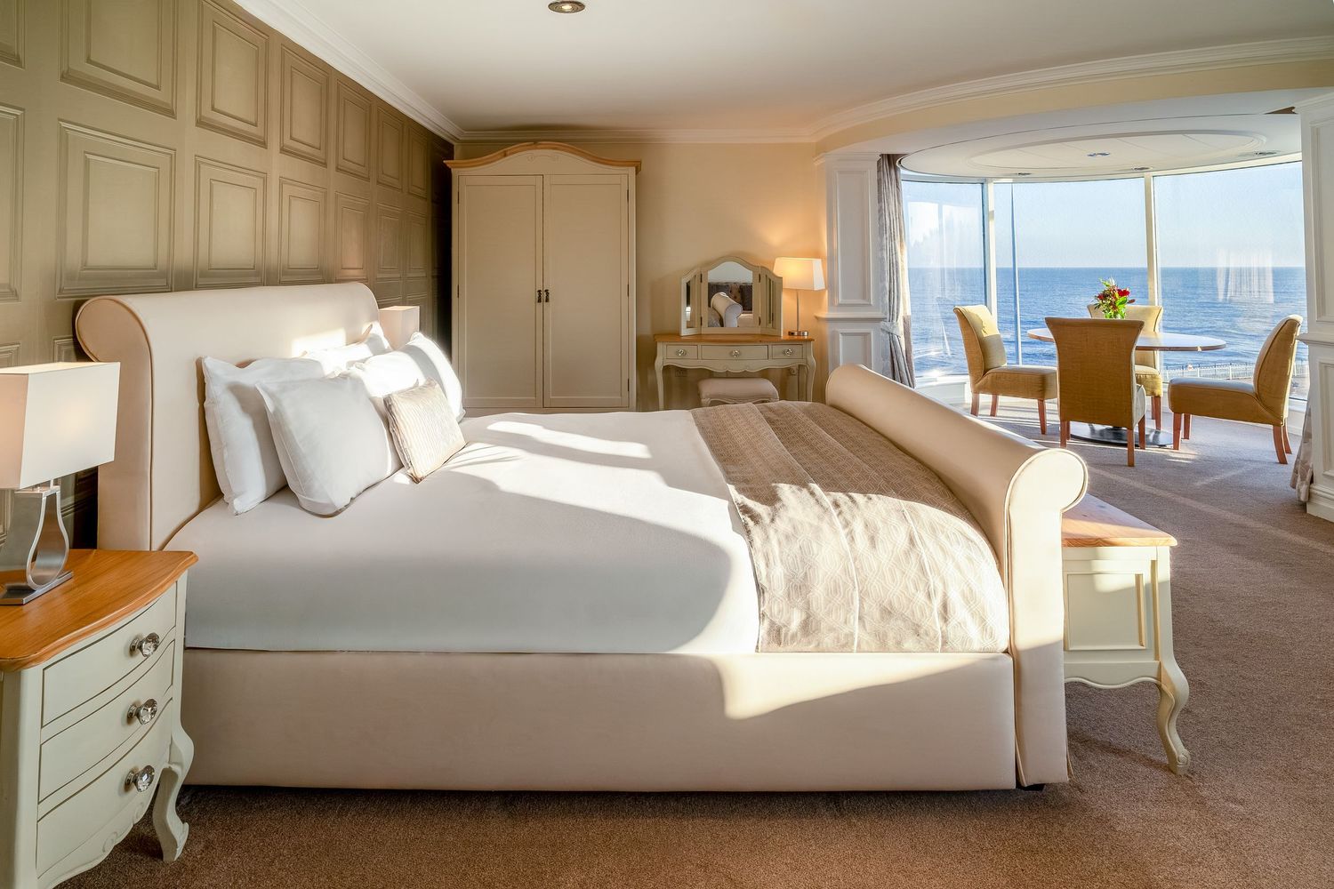 Elegant hotel bedroom with a plush white bed, ocean views, dining area, and warm natural light streaming through large windows.