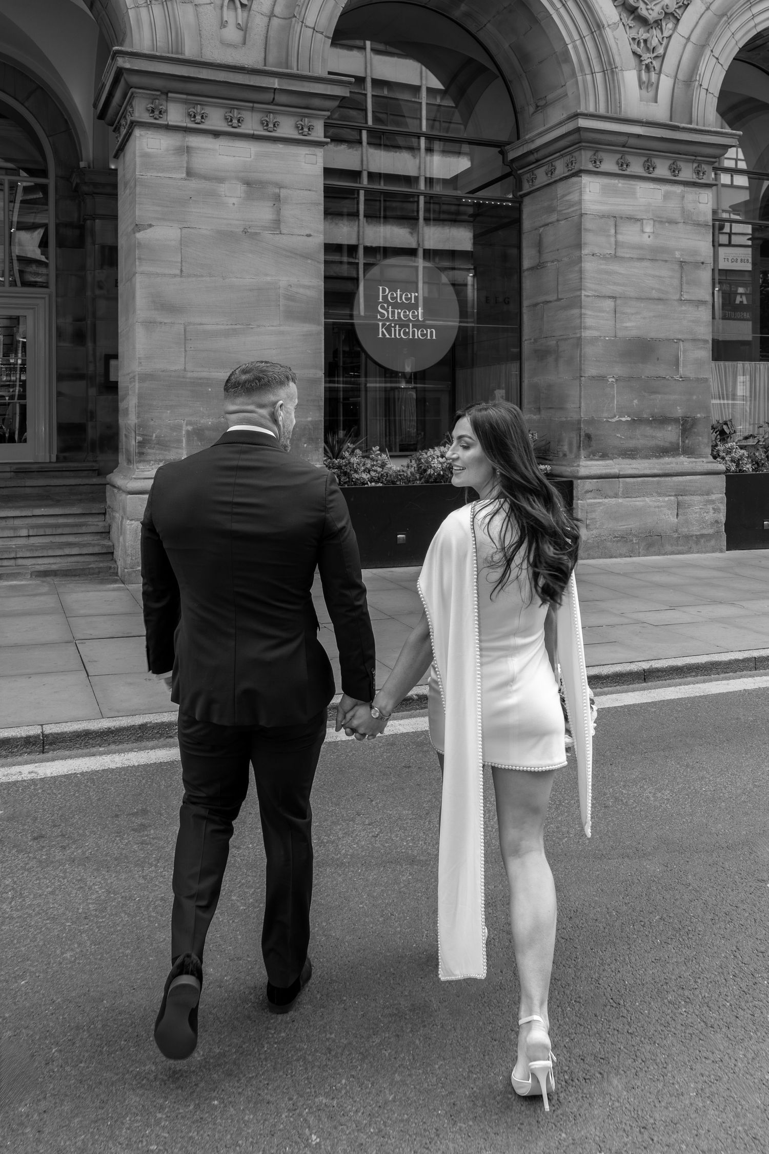 Black and white photo of a couple holding hands walking toward Peter Street Kitchen restaurant, woman in white mini dress.