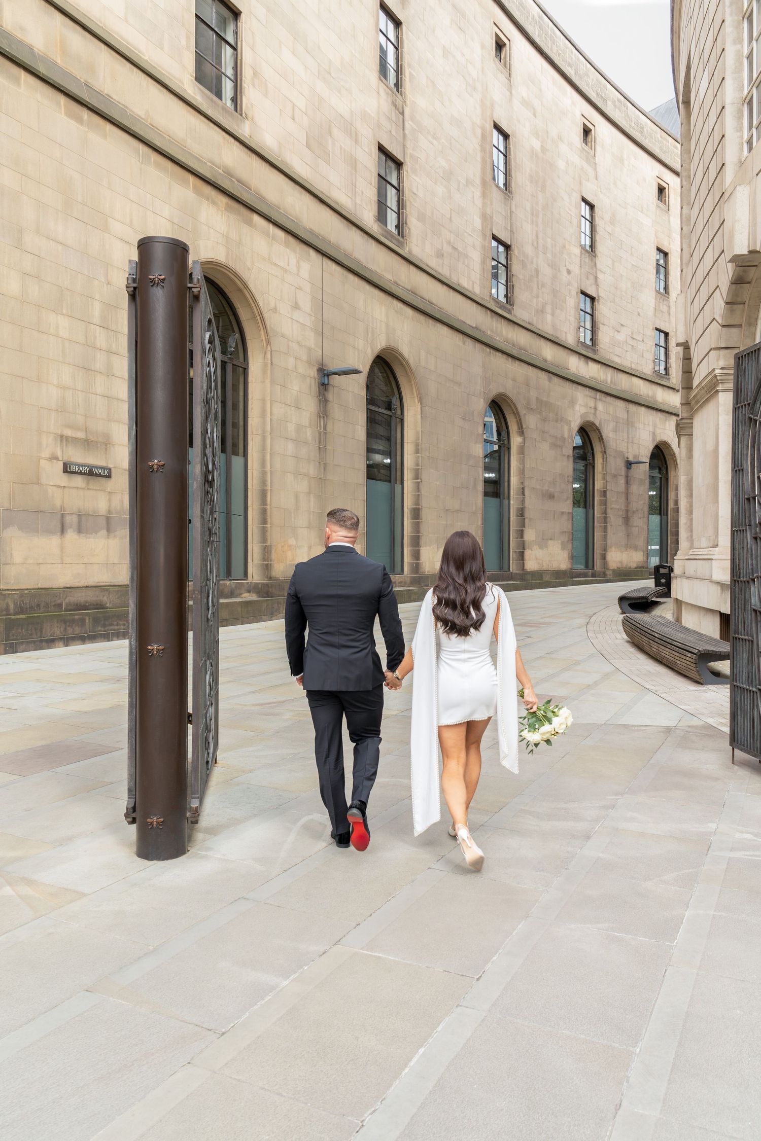 A bride and groom walk hand-in-hand along a curved stone building, she holds a bouquet in a short white dress.