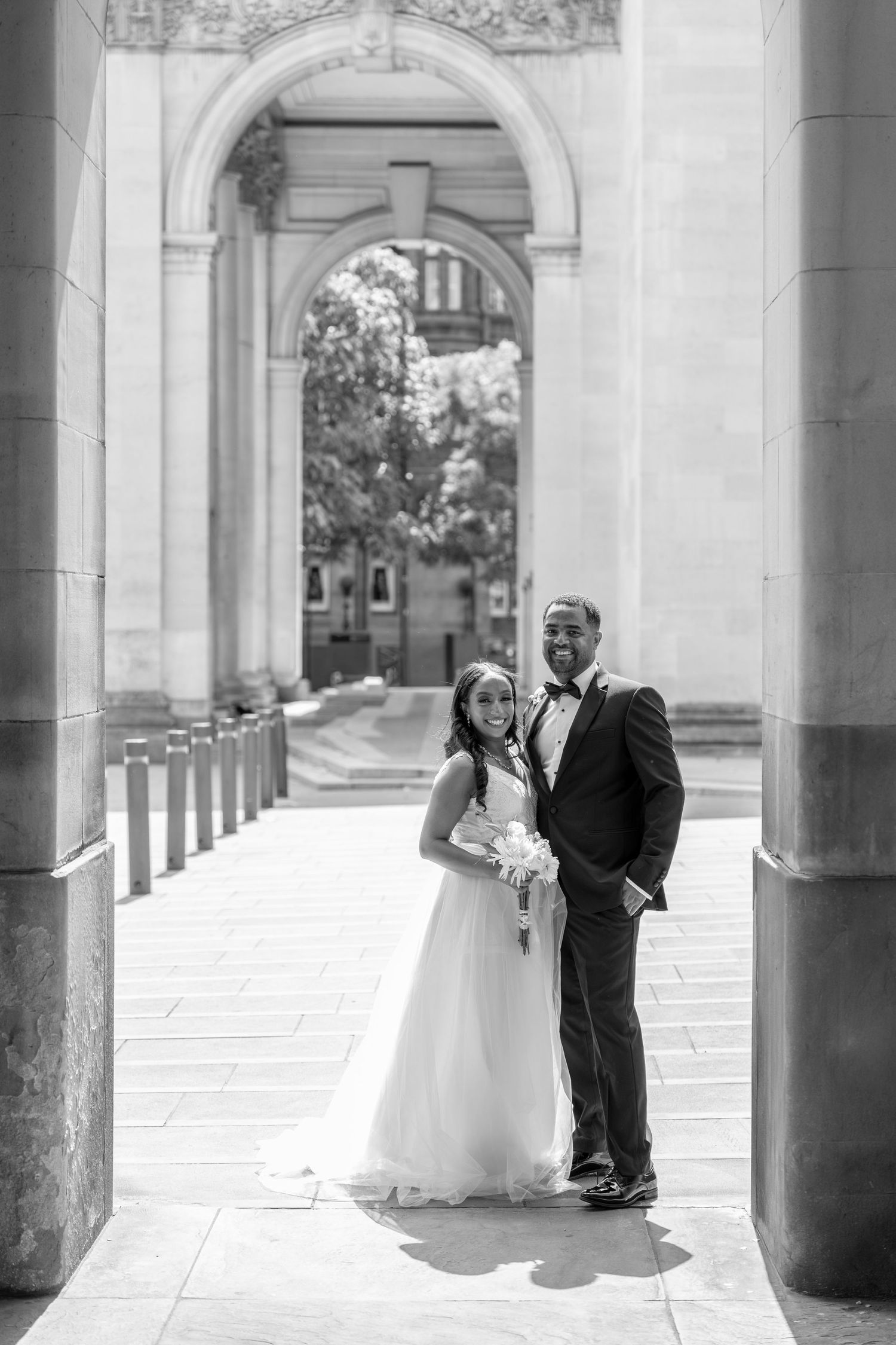 Black and white wedding photo of a bride and groom posing under a grand stone arch on a sunny day.