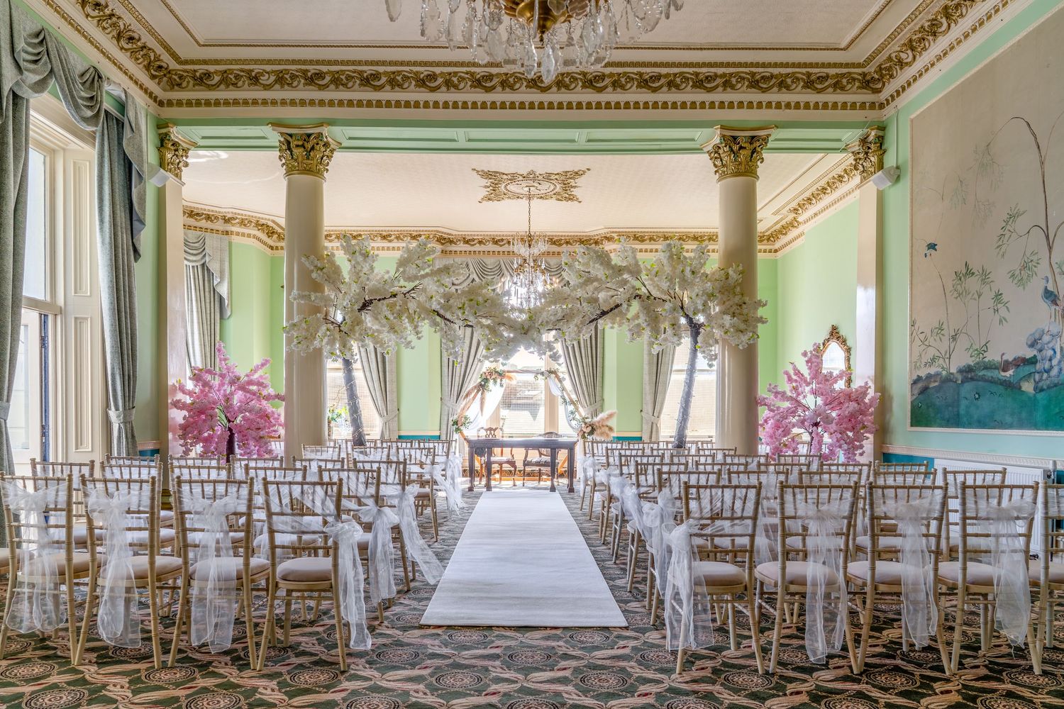 Elegant wedding ceremony venue with gold chairs, floral arch, pink centerpieces, and ornate green walls with gilded columns.