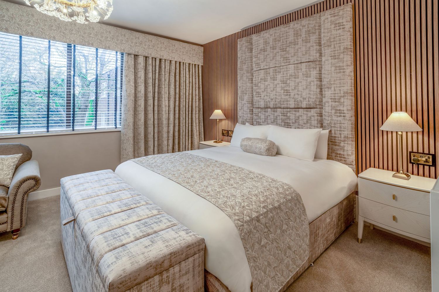 Elegant hotel bedroom with a large bed, neutral tones, padded headboard, bedside lamps, and natural light from a window.