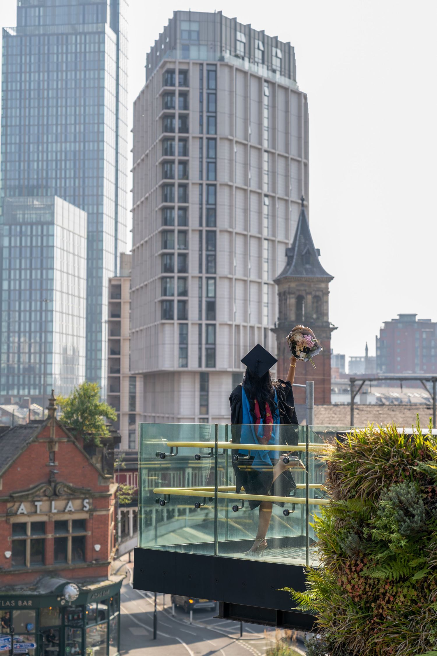 Graduate in cap and gown holds bouquet on rooftop balcony with Manchester city skyline and BBC tower behind.