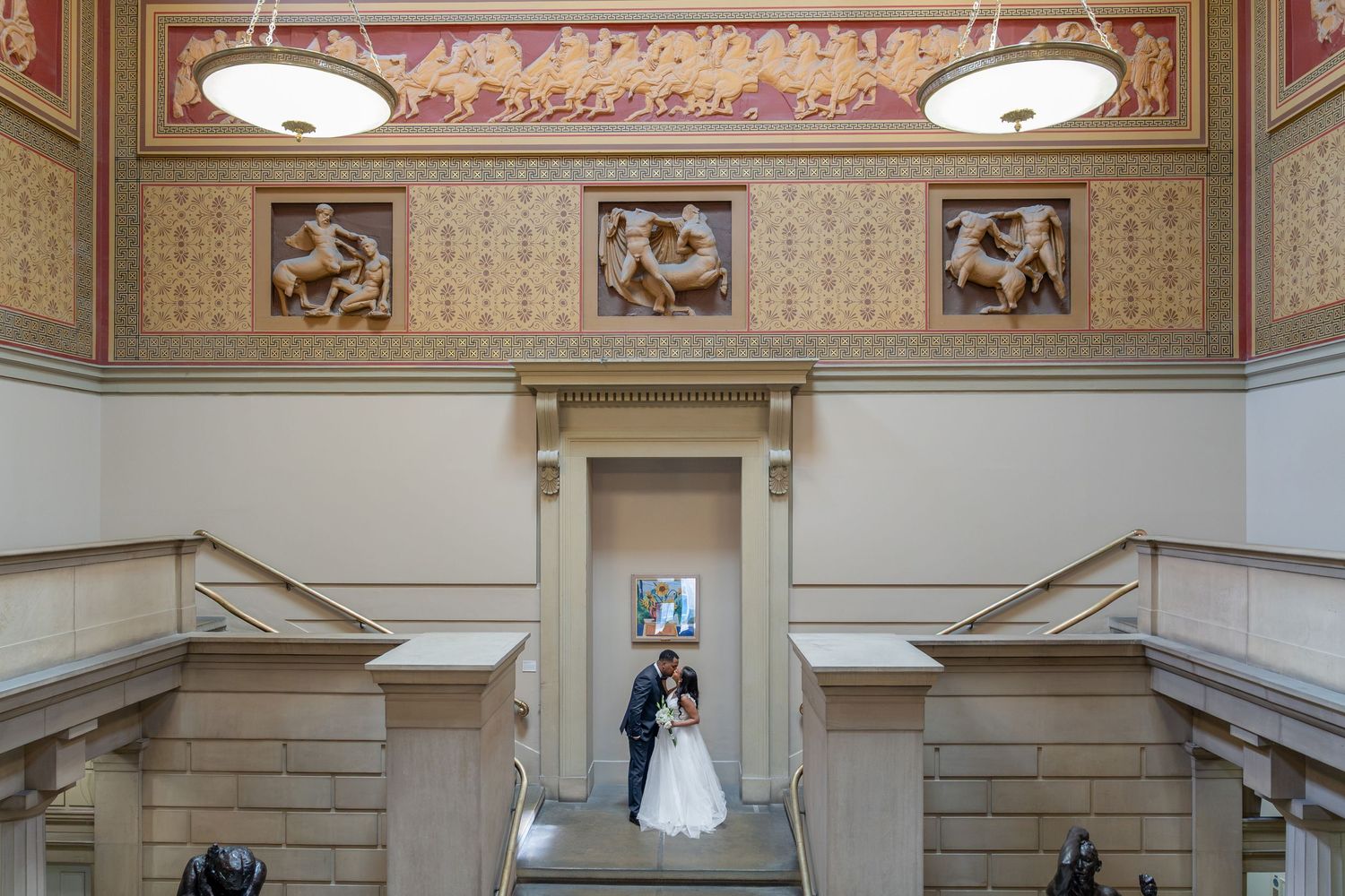 Bride and groom embrace in ornate museum staircase with classical murals and elegant architectural details.