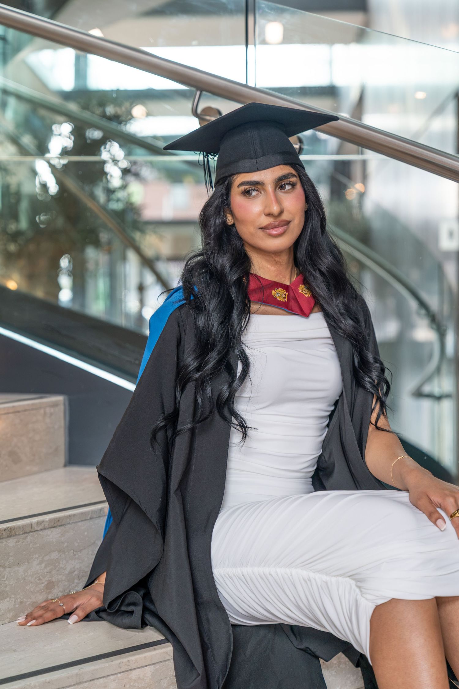 Young woman in graduation cap and gown over white dress, seated on stairs, celebrating academic achievement.