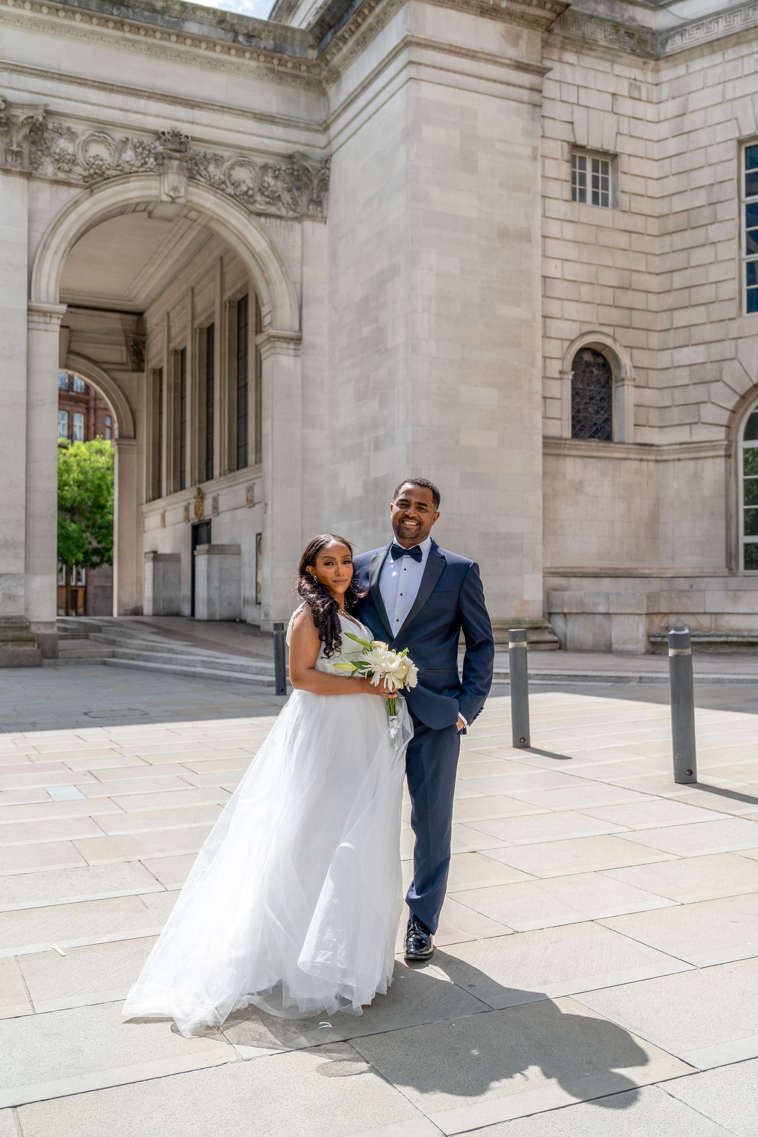 A bride in a white gown and groom in a navy suit pose outside a grand stone building on their wedding day.